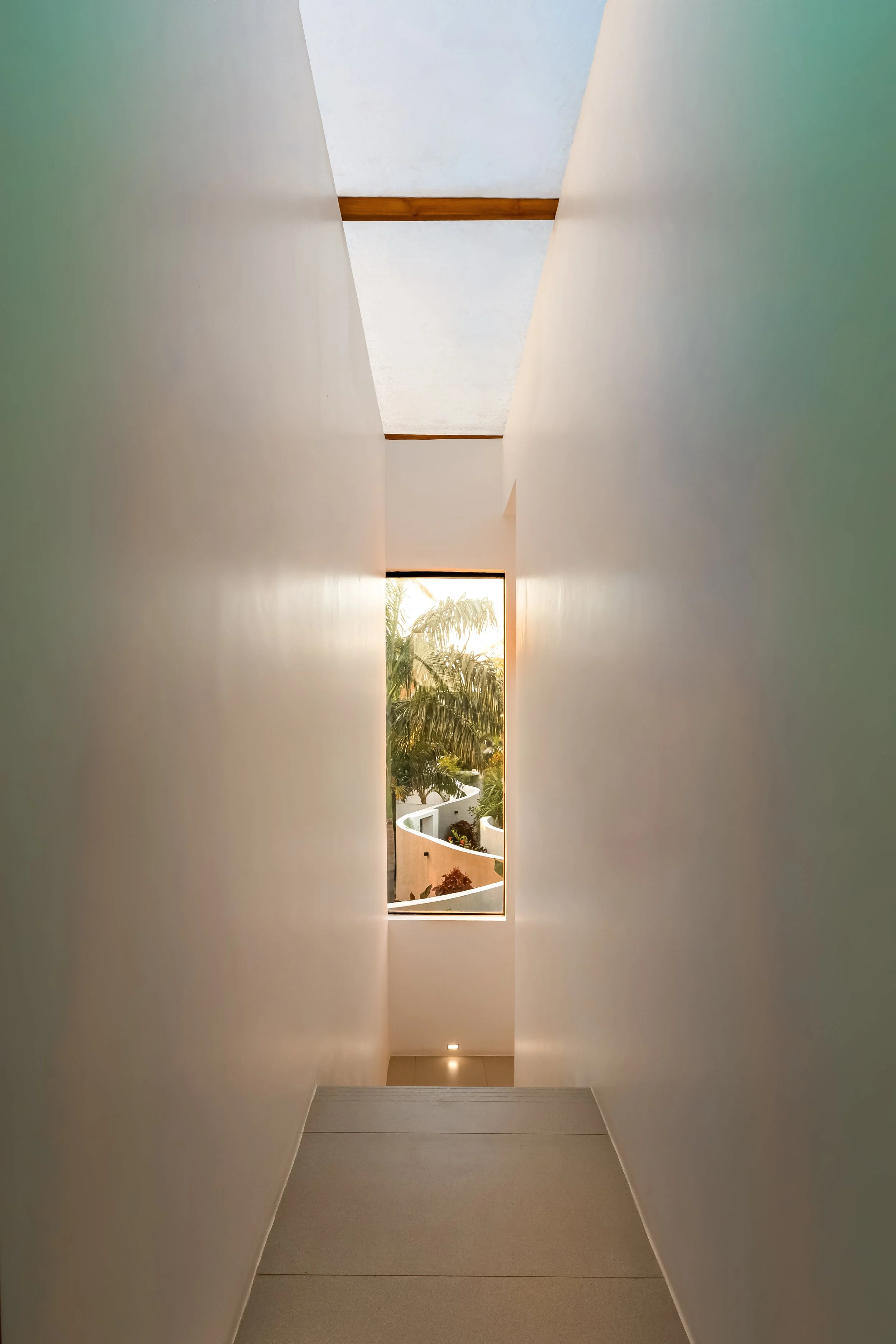View of a narrow corridor with white walls leading to a window with a tropical outdoor scene, including palm trees and sunlight.