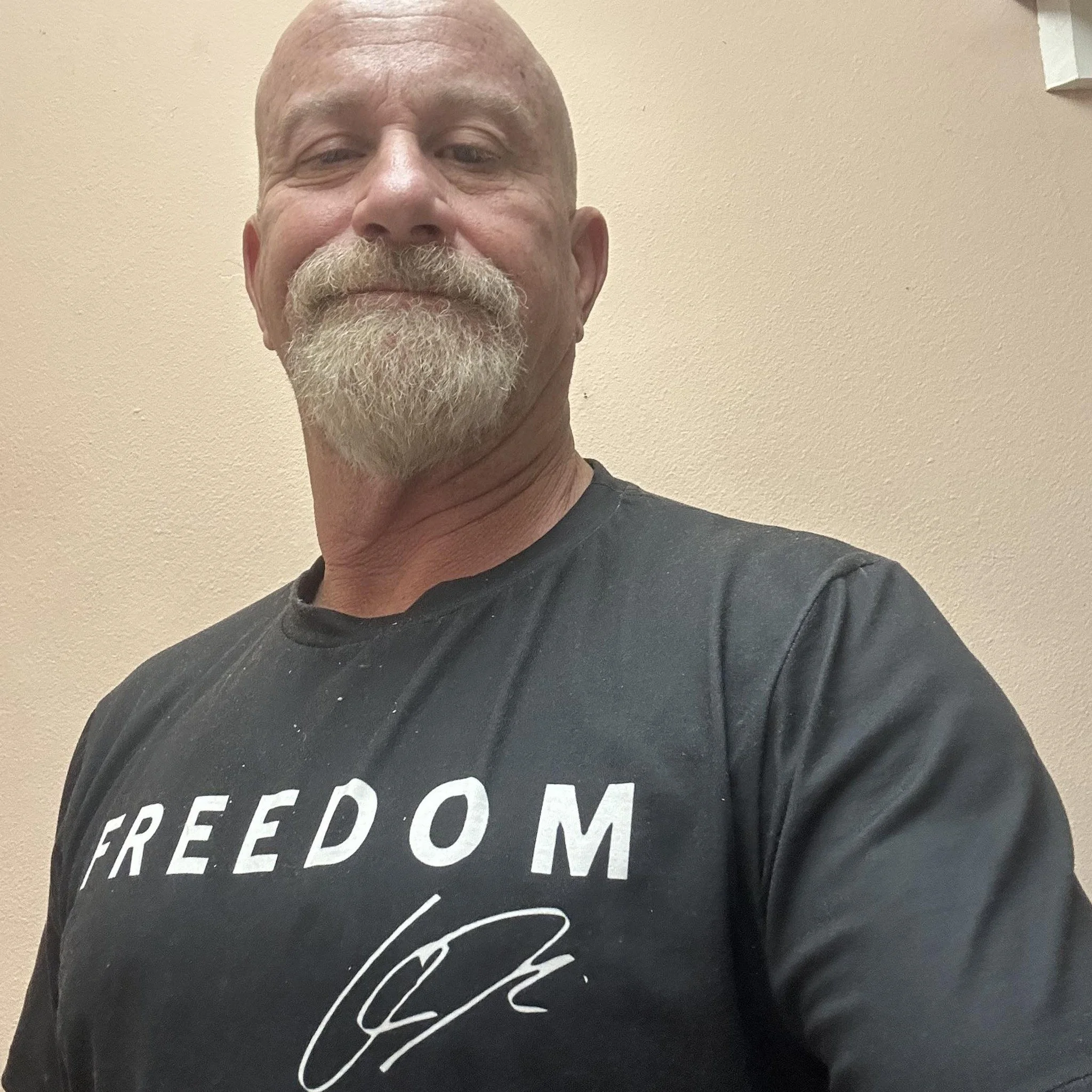 A middle-aged man with a bald head and a beard taking a selfie against a neutral wall. He is wearing a black T-shirt with the word 'FREEDOM' printed on it and a signature underneath.