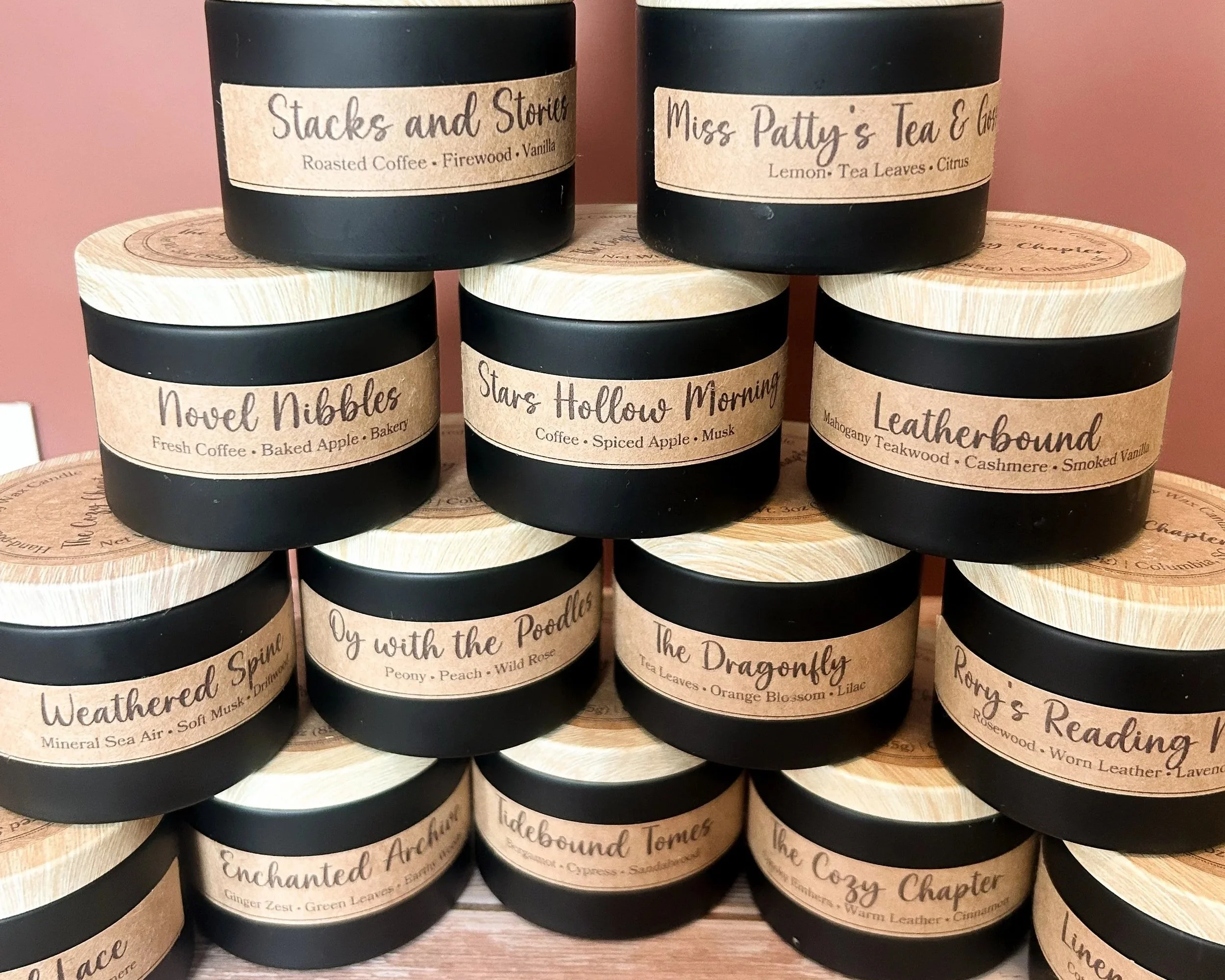 Stacked black jars with beige labels displaying different tea scents such as 'Stacks and Stories,' 'Miss Pattys Tea & Co.,' 'Novel Nibbles,' 'Stars Hollow Morning,' and others. The jars are placed on a round wooden display against a pink wall.