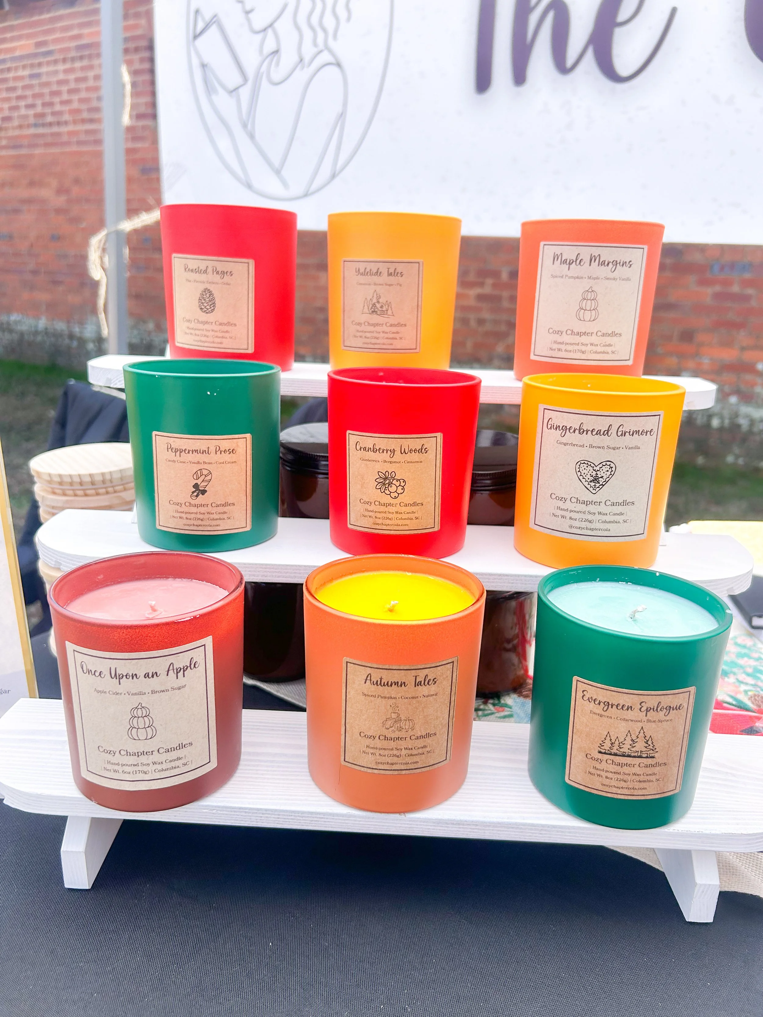 Colorful scented candles displayed on a tiered white stand at an outdoor event, with a backdrop of a brick wall and a banner.