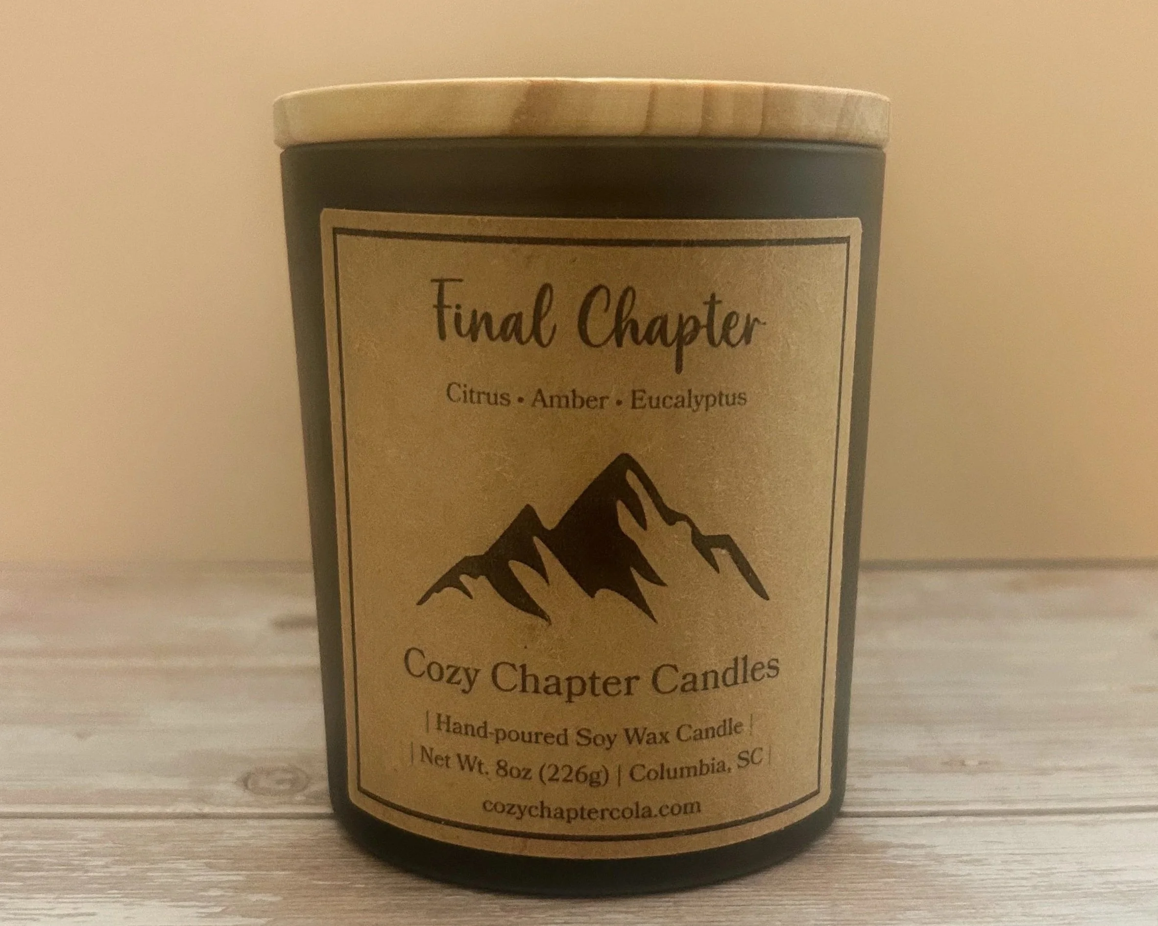A black candle jar with a beige lid labeled 'Final Chapter' and scents like Citrus, Amber, and Eucalyptus. The label also describes it as a cozy chapter candle, hand-poured soy wax, weighing 8oz, from Columbia, SC.