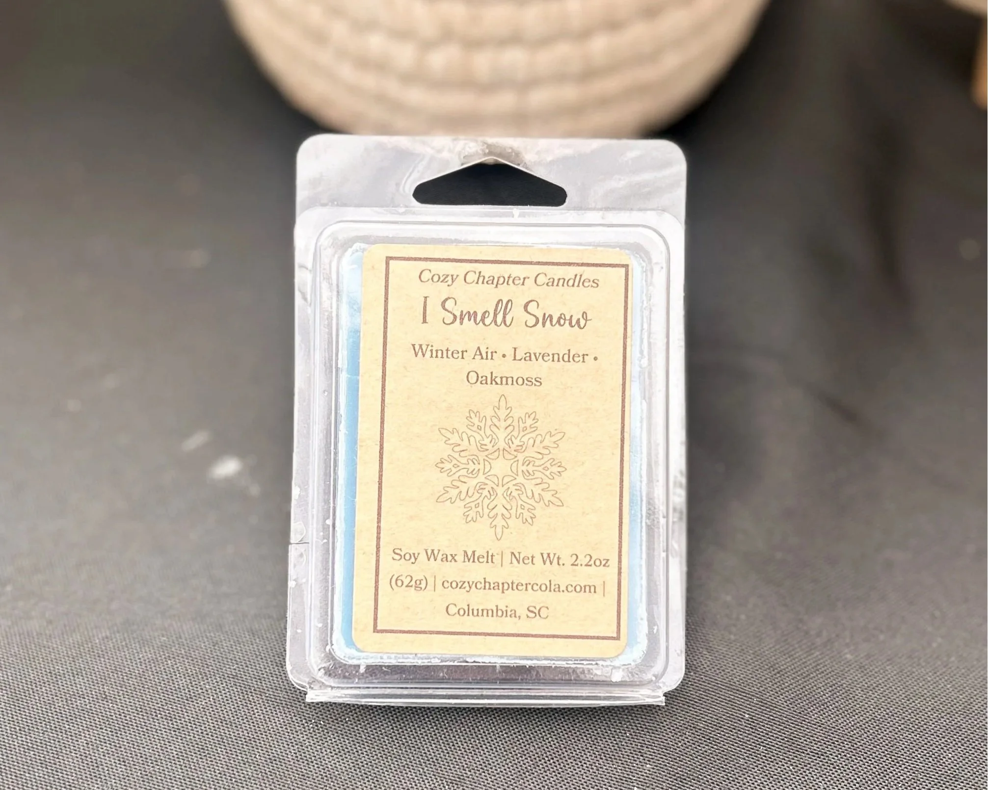 Close-up of a package of a Cozy Chapter Wax Melt, named 'I Smell Snow,' with a snowflake illustration and botanical ingredients listed, resting on a dark surface. Scent is inspired by a popular 90s TV show and a part of the collection, Gazebo Glow.