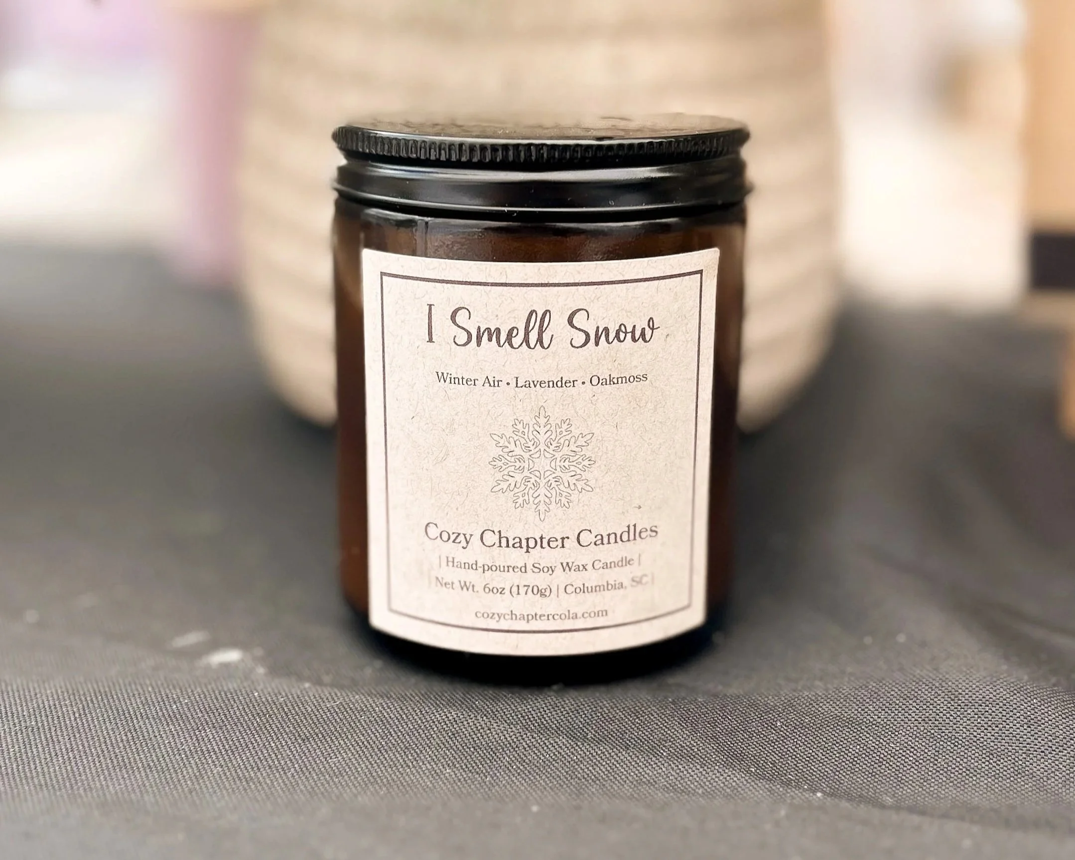 A close-up of a brown candle jar with a white label reading "I Smell Snow" and scent notes: Winter Air, Lavender, Oakmoss. The jar has a black lid and is placed on a grey fabric surface, with blurred background.