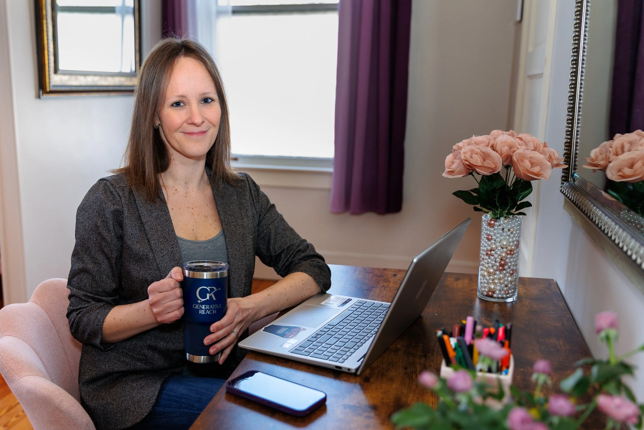 Janina Grayeb owner of Generative Reach sitting at a desk with her computer, phone and holding a coffee tumbler