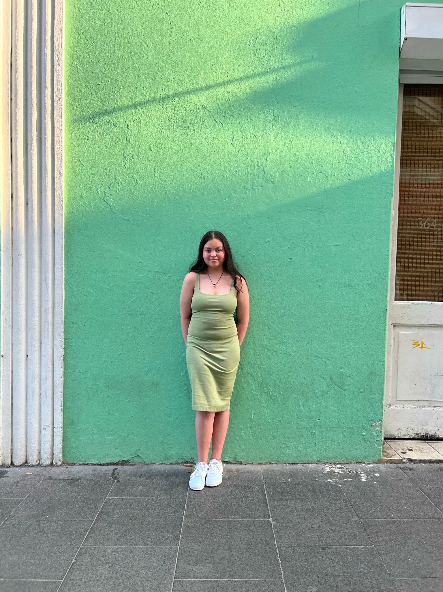 Image of Leah wearing a green dress and white shoes against a green wall.