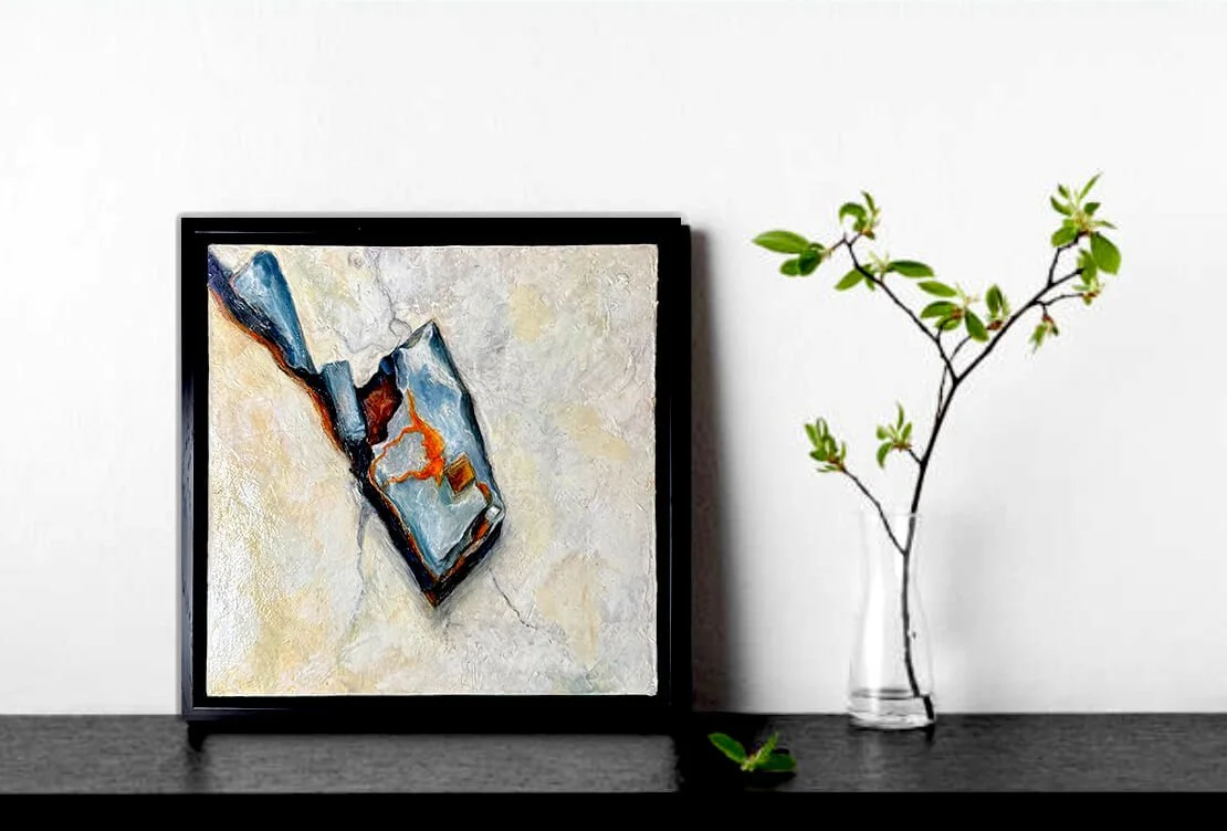 An abstract painting in a black frame leaning against a white wall, with a small branch with green leaves in a clear vase beside it on a dark surface. Small artwork 12x12
