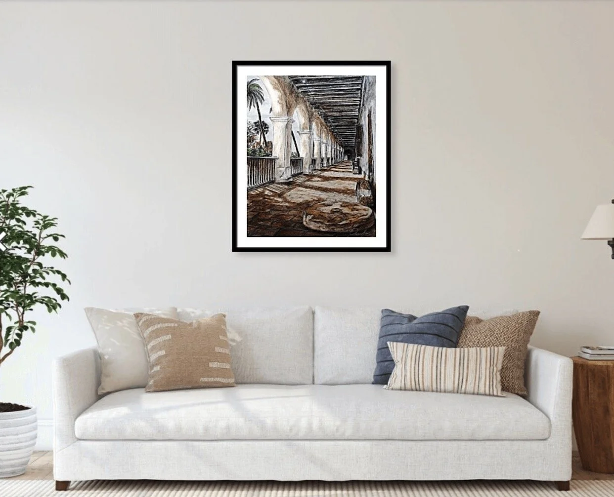 Living room with white sofa, beige and blue pillows, a side table with books, a wall lamp, a potted plant, and a framed painting of a covered porch with arches and palm trees.