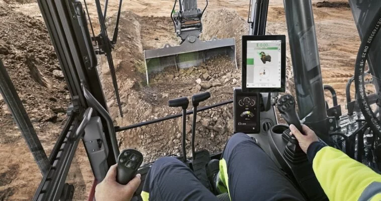 Inside a construction excavator cabin with a person operating the machinery, view of the dirt excavation site outside.