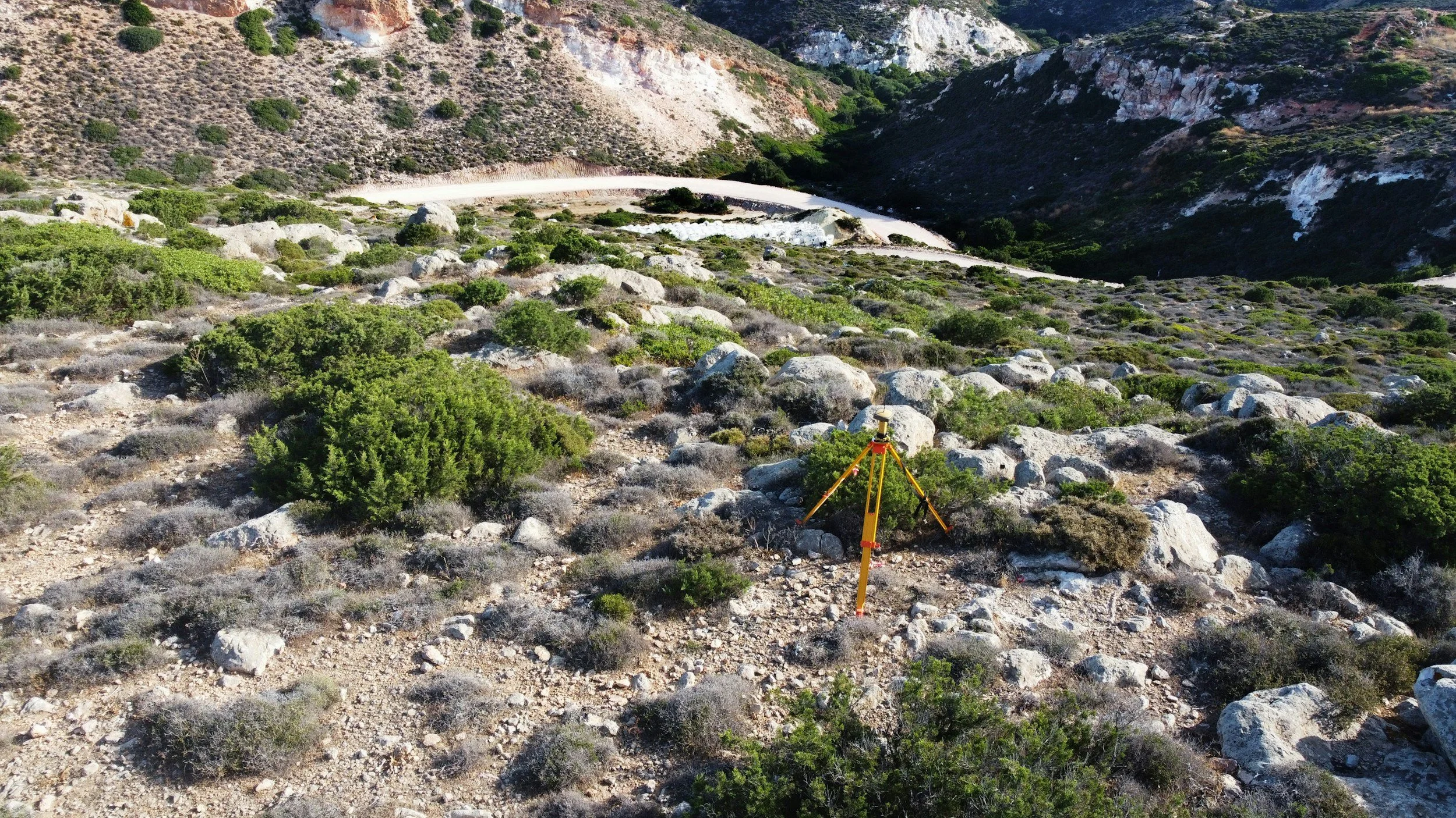 A surveyor tripod set up in a rocky, shrub-covered mountainside with a winding dirt road visible in the distance.