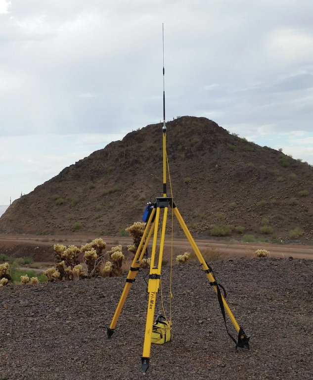 Surveying tripod with yellow legs set up in a desert landscape, with cacti and a hill in the background under a cloudy sky.