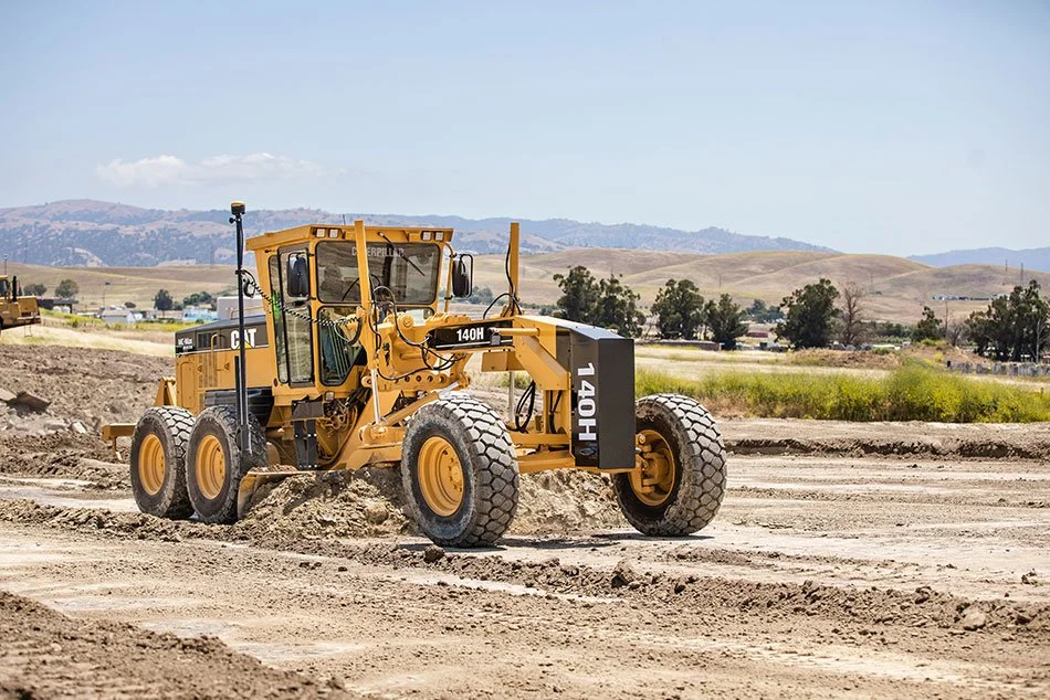 A yellow motor grader working on a construction site with a mountainous landscape in the background