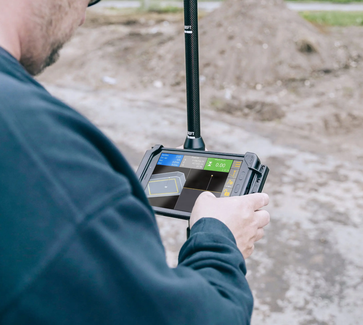 A person in a dark jacket holding a rugged tablet device, which displays design or surveying software, outdoors with a construction site or dirt ground in the background.