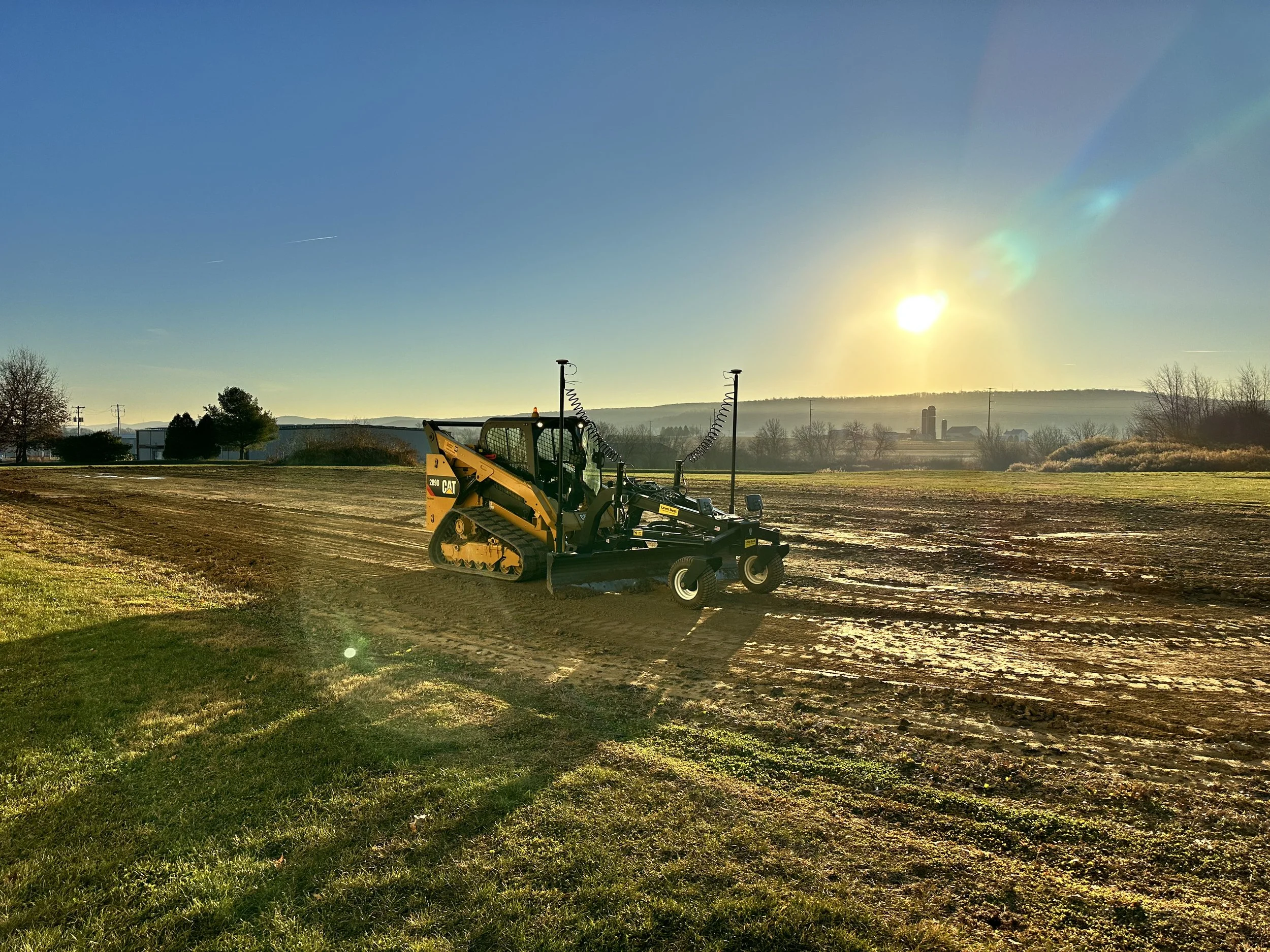 A small yellow and black Caterpillar compact track loader sits on a plowed field at sunset.