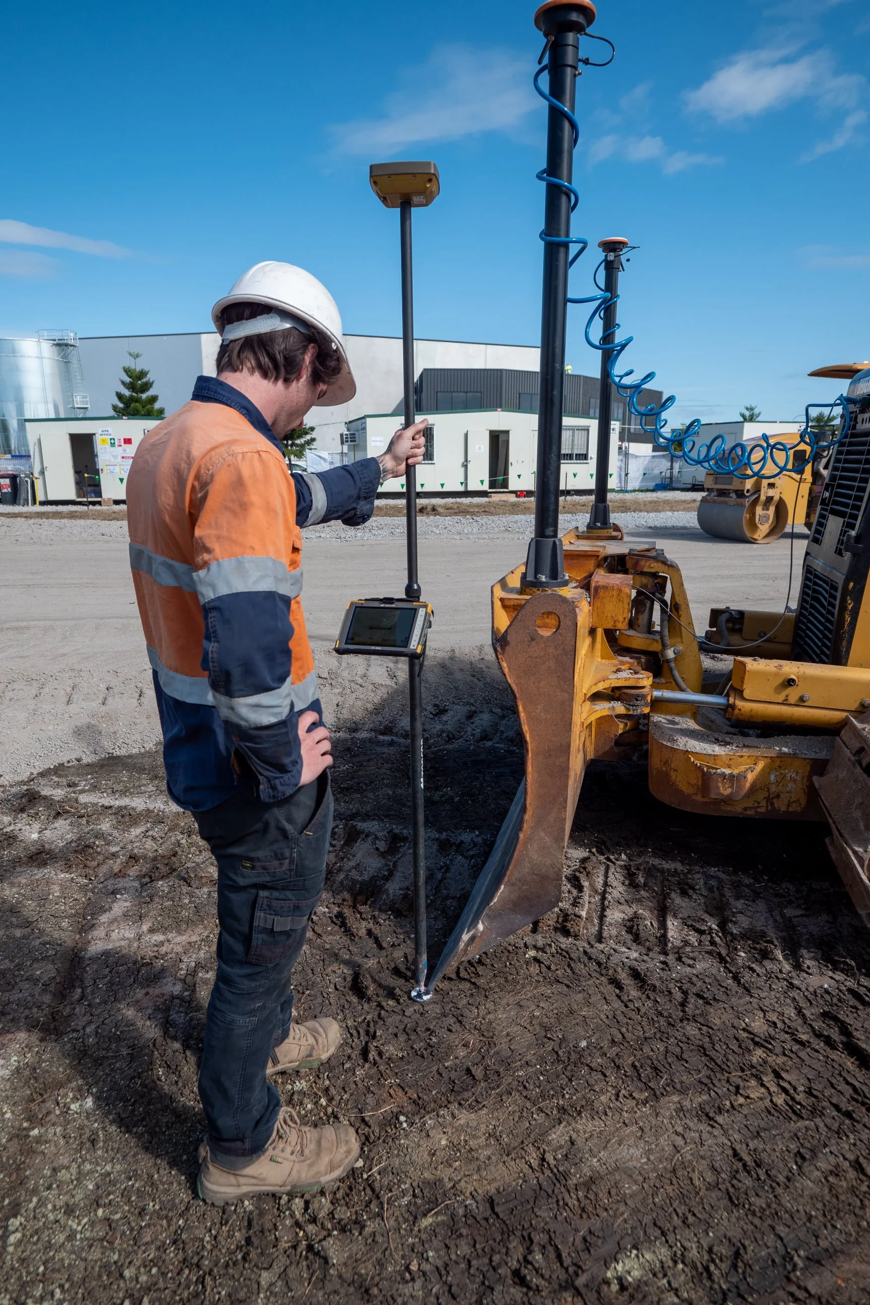 Construction worker standing beside a piece of equipment on a construction site, inspecting or adjusting it, with a blue sky and industrial buildings in the background.
