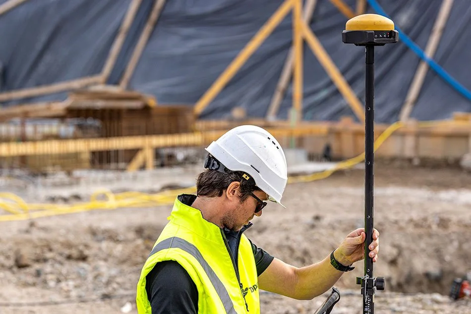 A construction worker wearing a white safety helmet and a bright yellow safety vest at a construction site, using a surveying device mounted on a tall pole to measure the terrain.