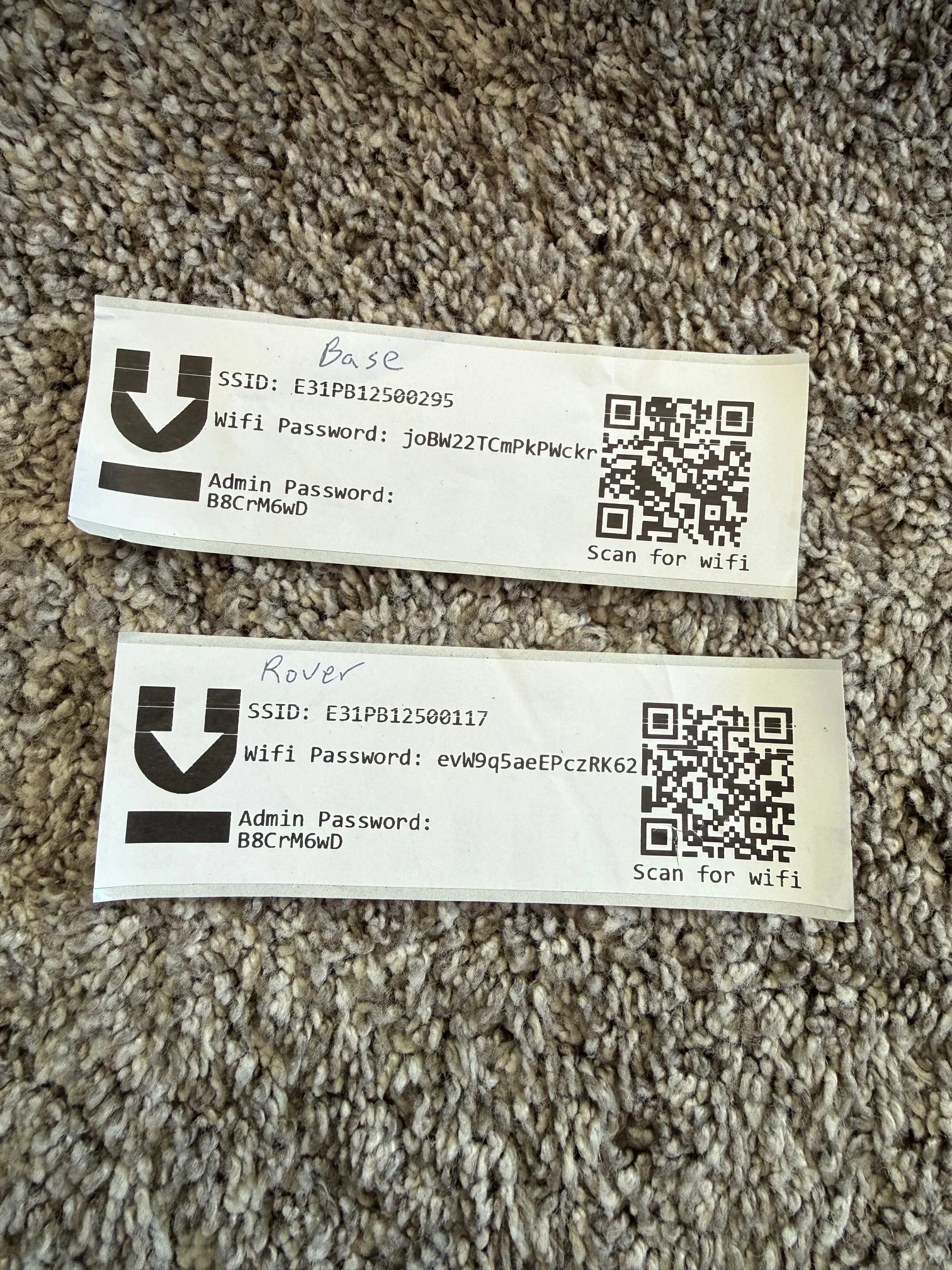 Two WiFi access point labels on a carpet. The top label is for a WiFi network named 'Base' with password 'joBw22TCmPkPWckr' and admin password 'B8CrM6wD.' The bottom label is for a WiFi network named 'Rover' with password 'evW9q5aeEPczRK62' and the same admin password. Each label has a QR code for scanning.