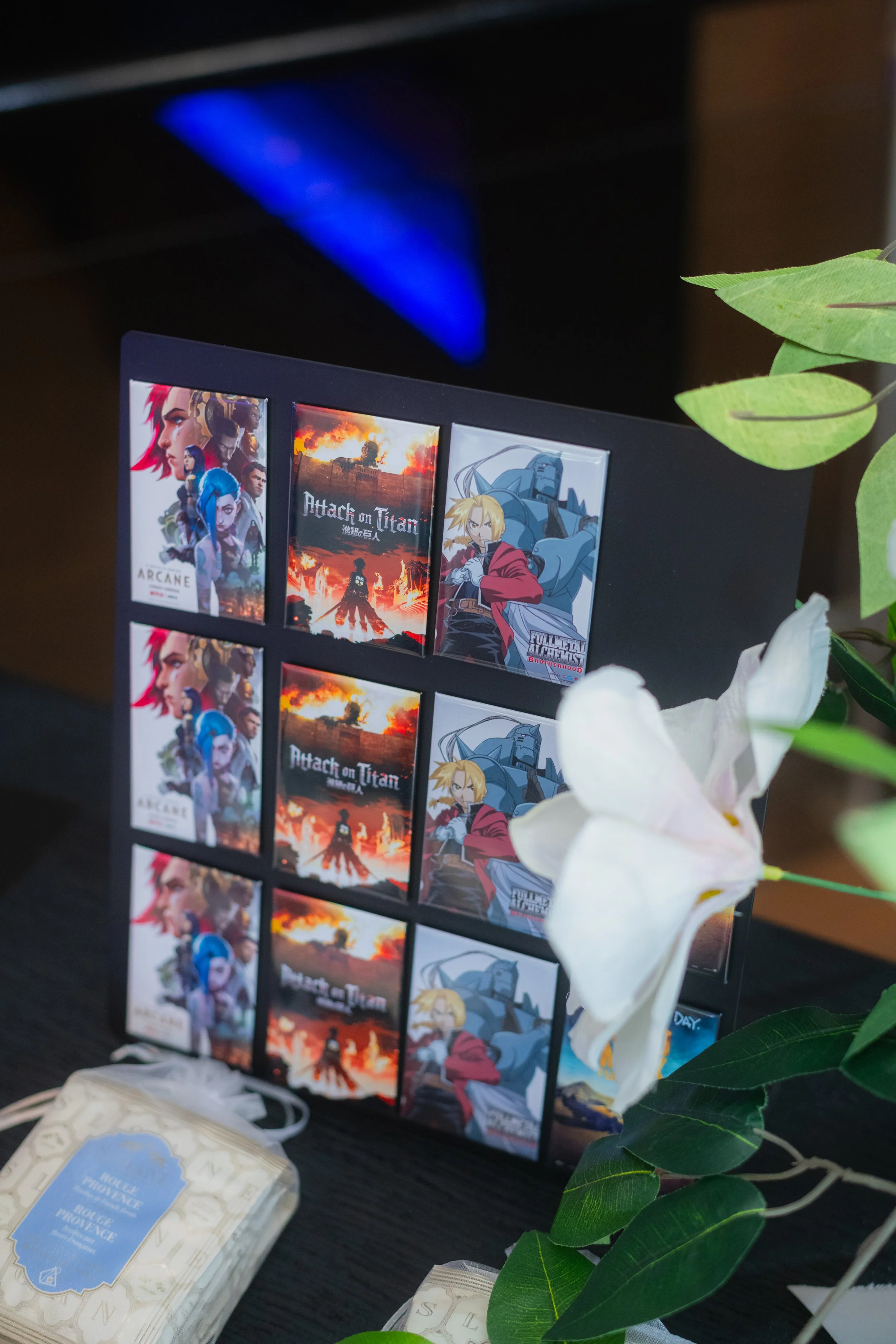 A display of several stickers on a black surface, featuring characters from anime and games, with part of a greenery and white flowers visible in the foreground and a blurred blue-lit background.