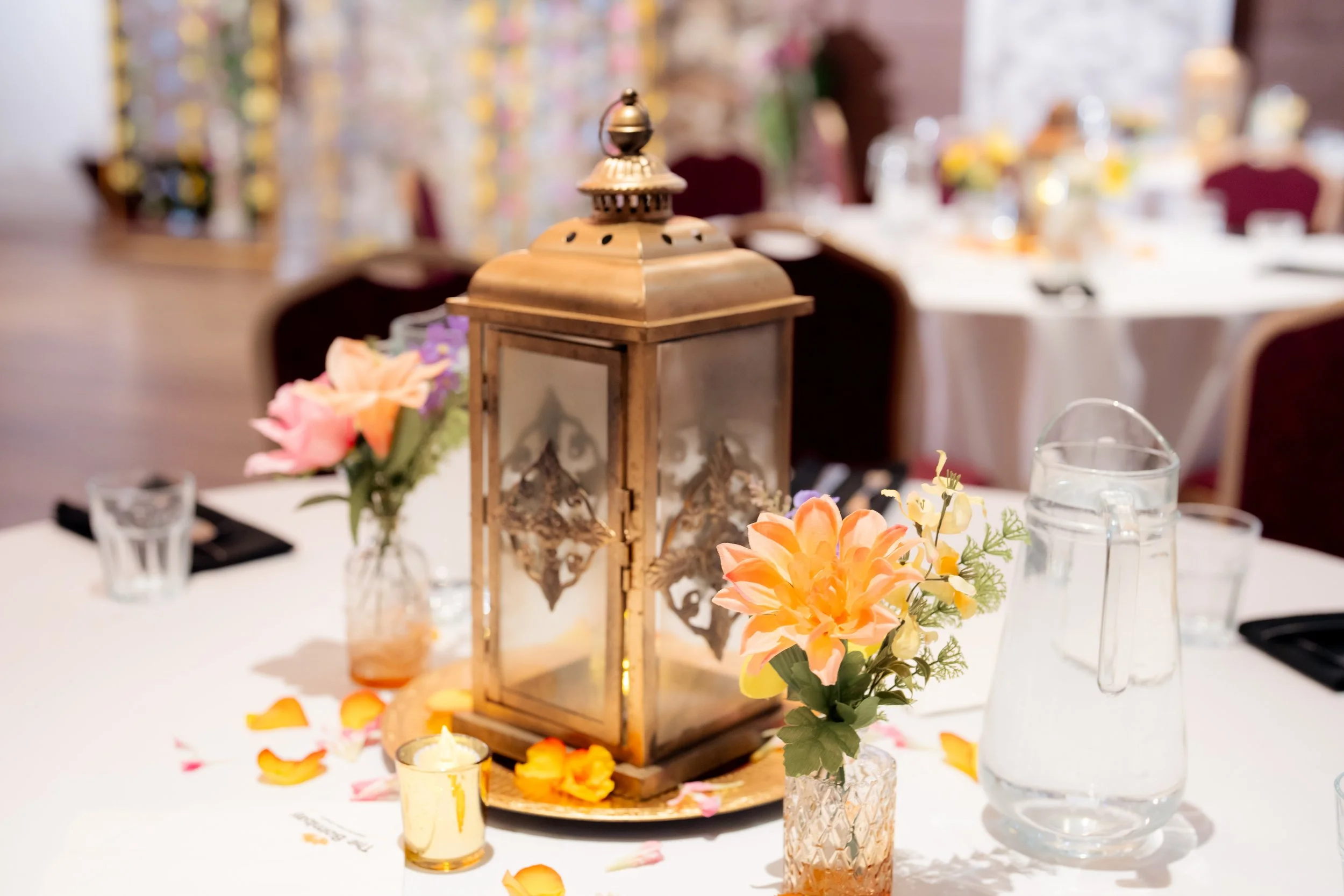 A decorative table centerpiece featuring a wooden lantern surrounded by colorful flowers, candles, and scattered petals at an elegant event setting.