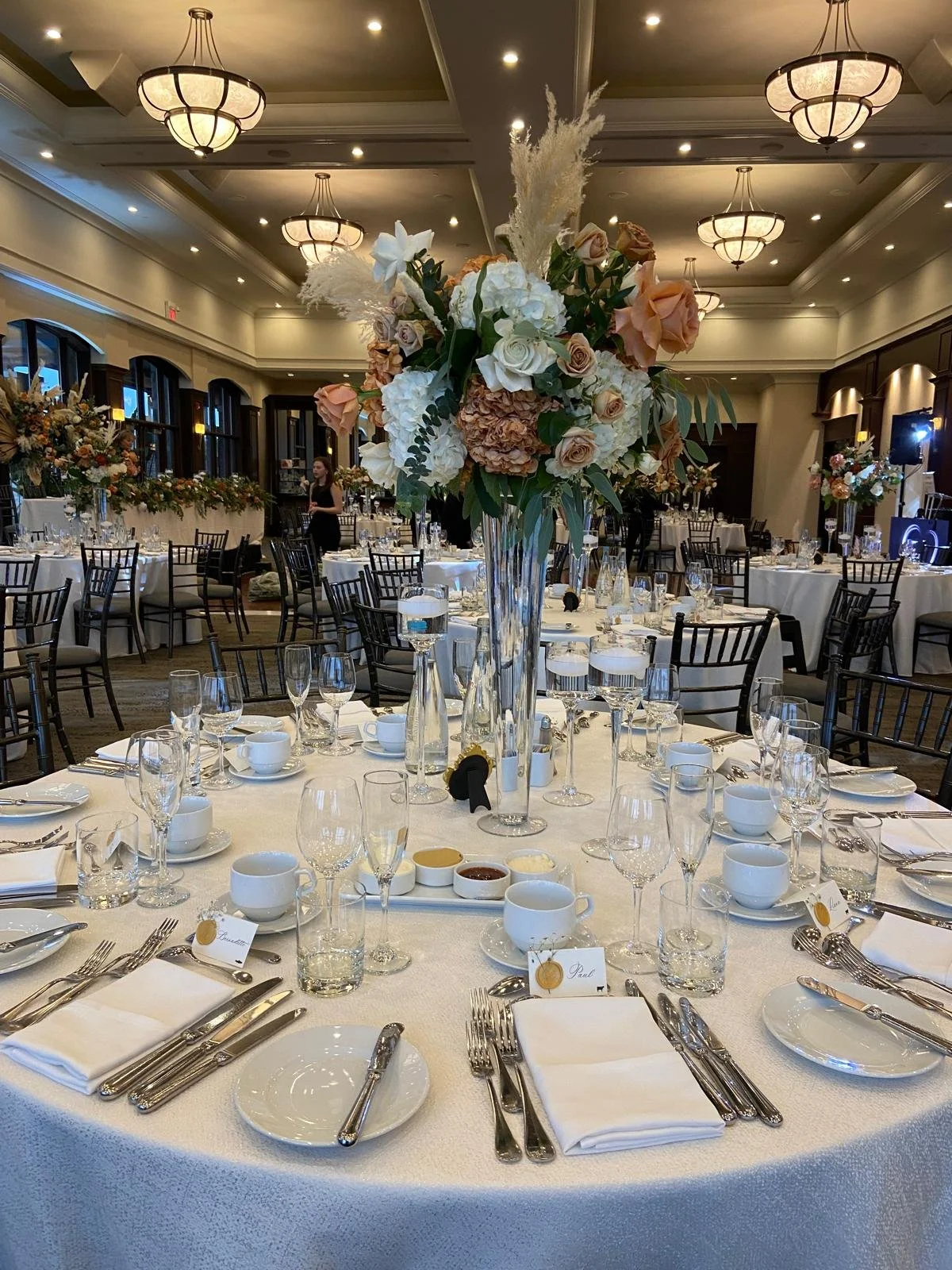 Elegant banquet hall decorated for a wedding or formal event with round tables covered in white tablecloths. Each table features a tall floral centerpiece with white, peach, and blush roses, hydrangeas, and greenery. Place settings include white napk