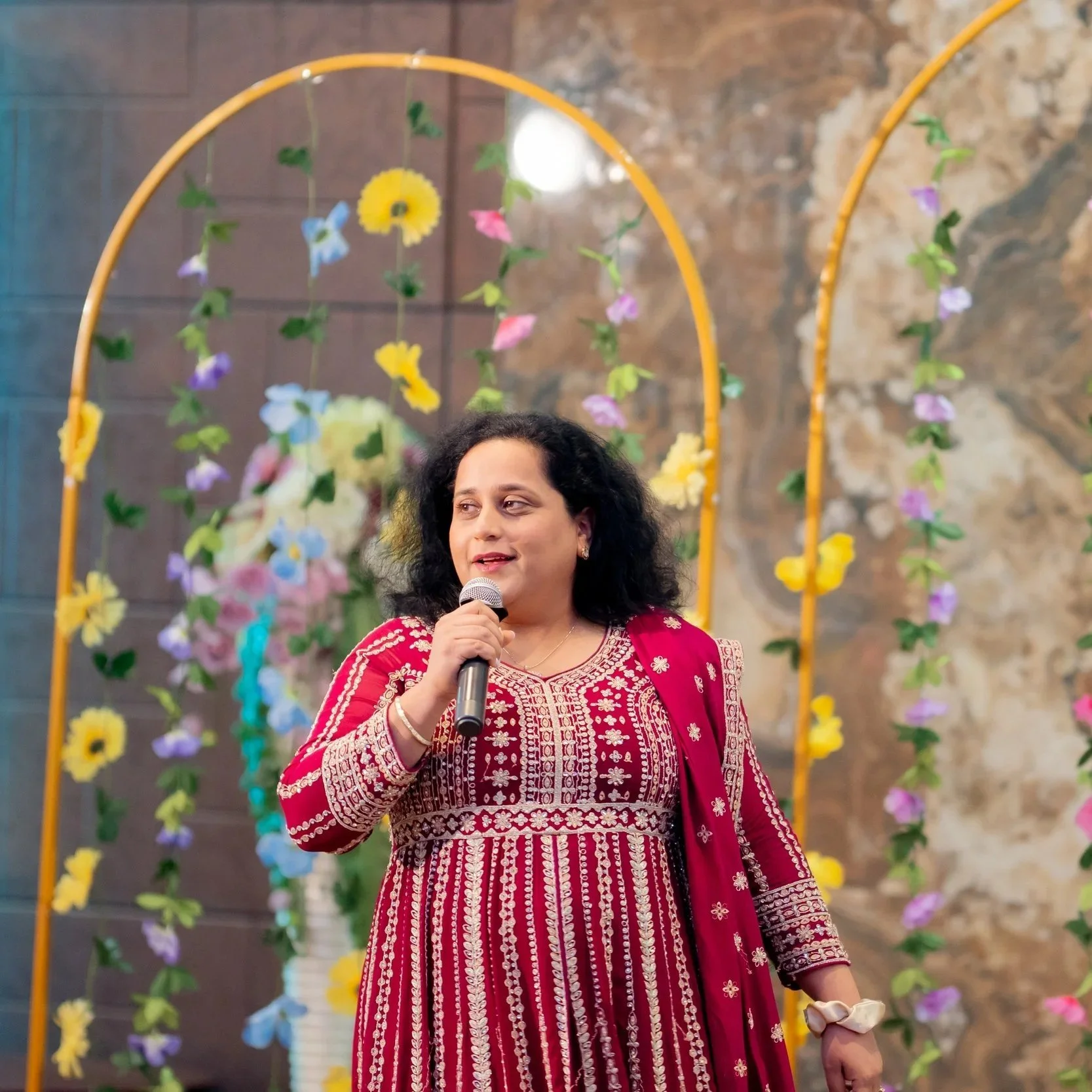 A woman wearing a red and white traditional dress is holding a microphone and speaking in front of a decorative floral arch with yellow, purple, and blue flowers. The background appears to be an indoor setting with a stone wall.