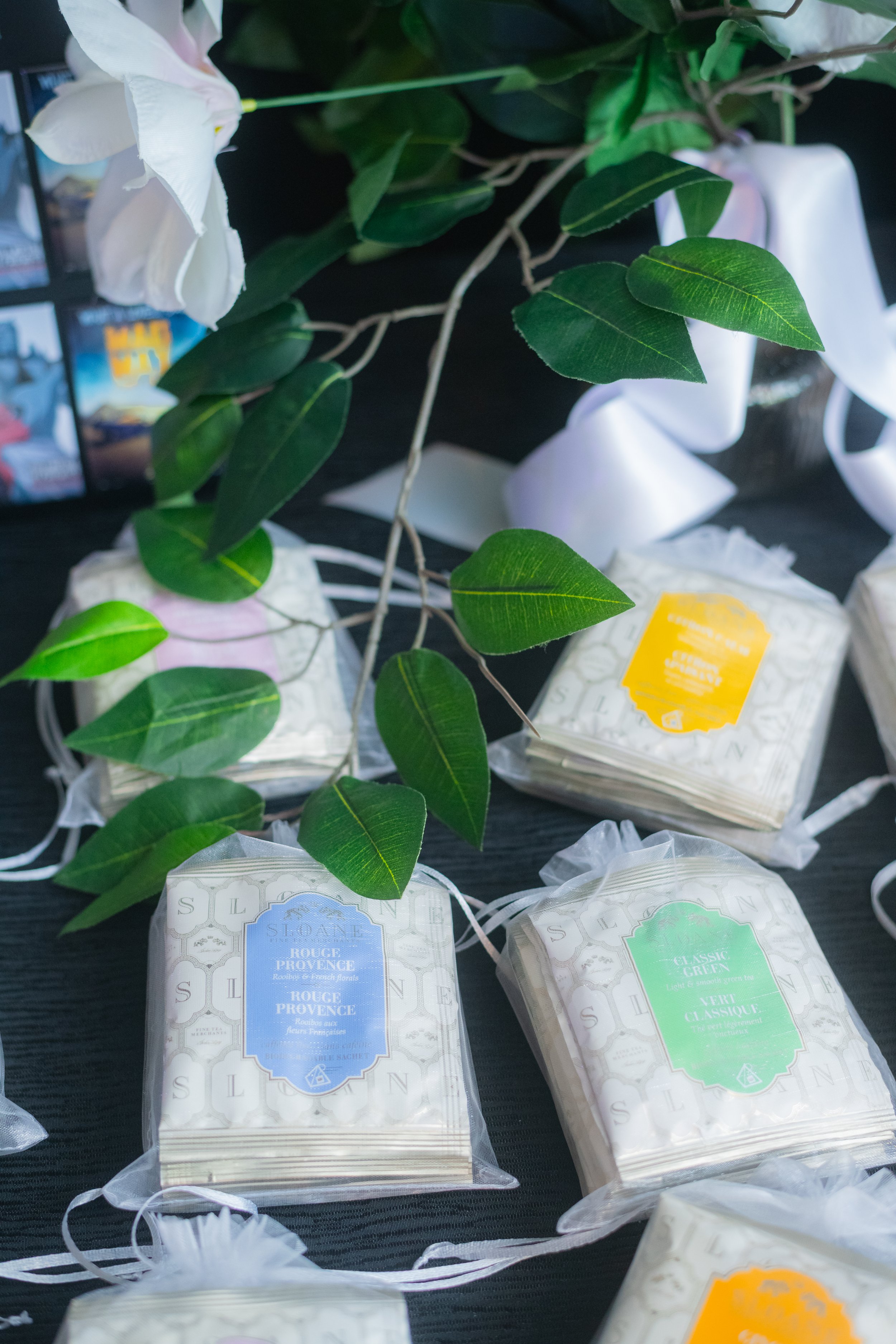 Bundles of packaged tea and artificial green leaves on a dark surface, with a blurred background showing a monitor and other objects.
