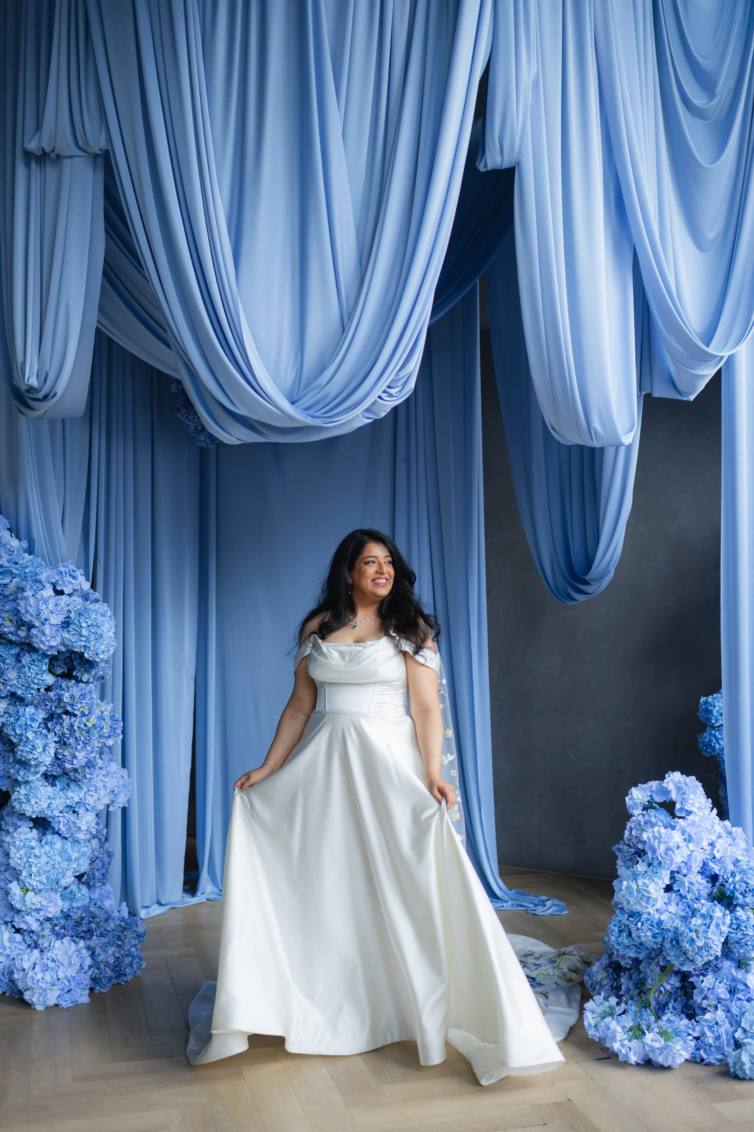 A woman in a white wedding dress standing in front of a blue floral archway with draped blue fabric.