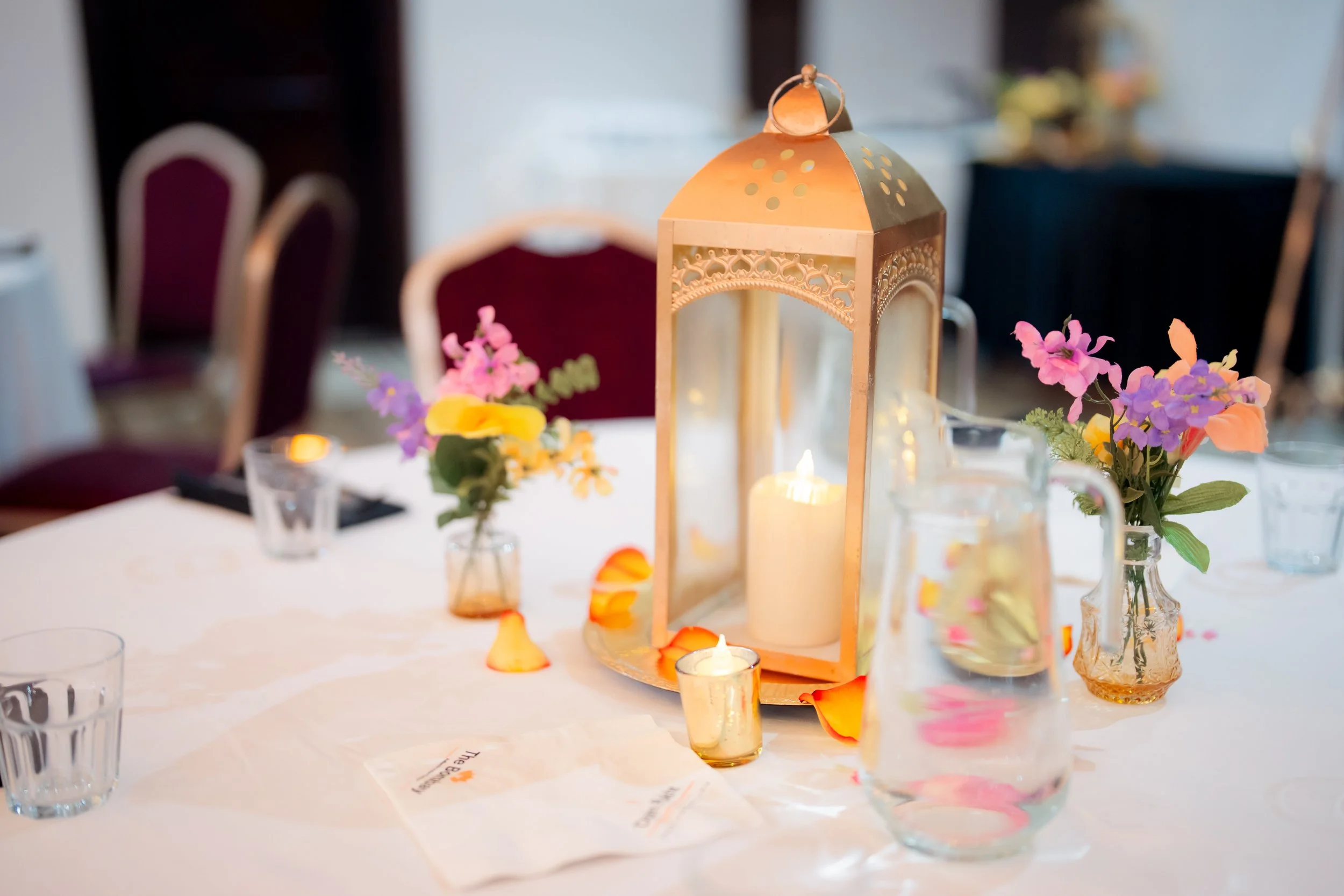 A centerpiece on a banquet table featuring a wooden lantern with decorative cutouts, surrounded by small glass vases with colorful flowers and lit candles, set in a banquet hall with red chairs.