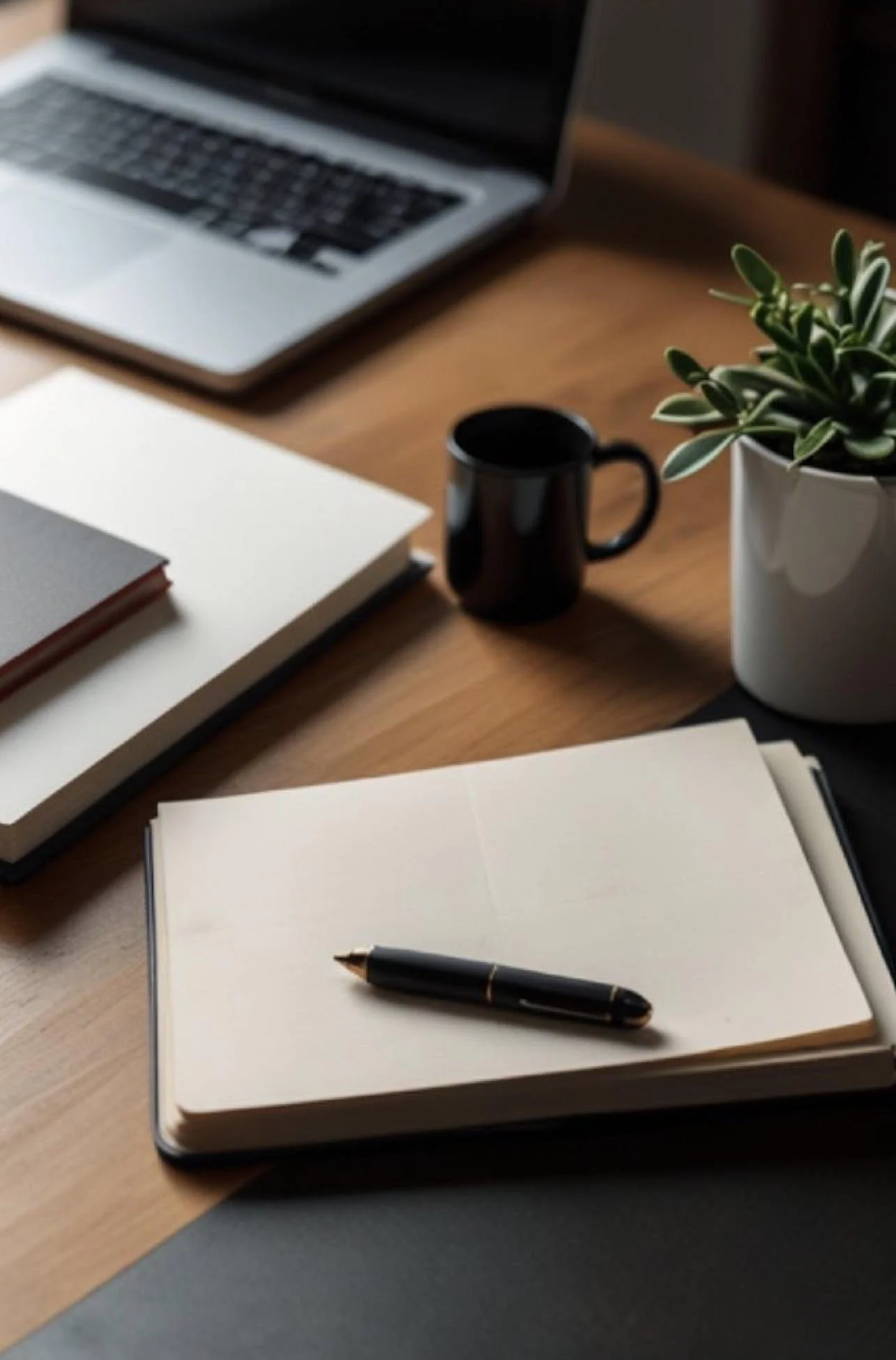 Person typing on a silver laptop with a black screen, on a dark table, with a blurred plant in the background.
