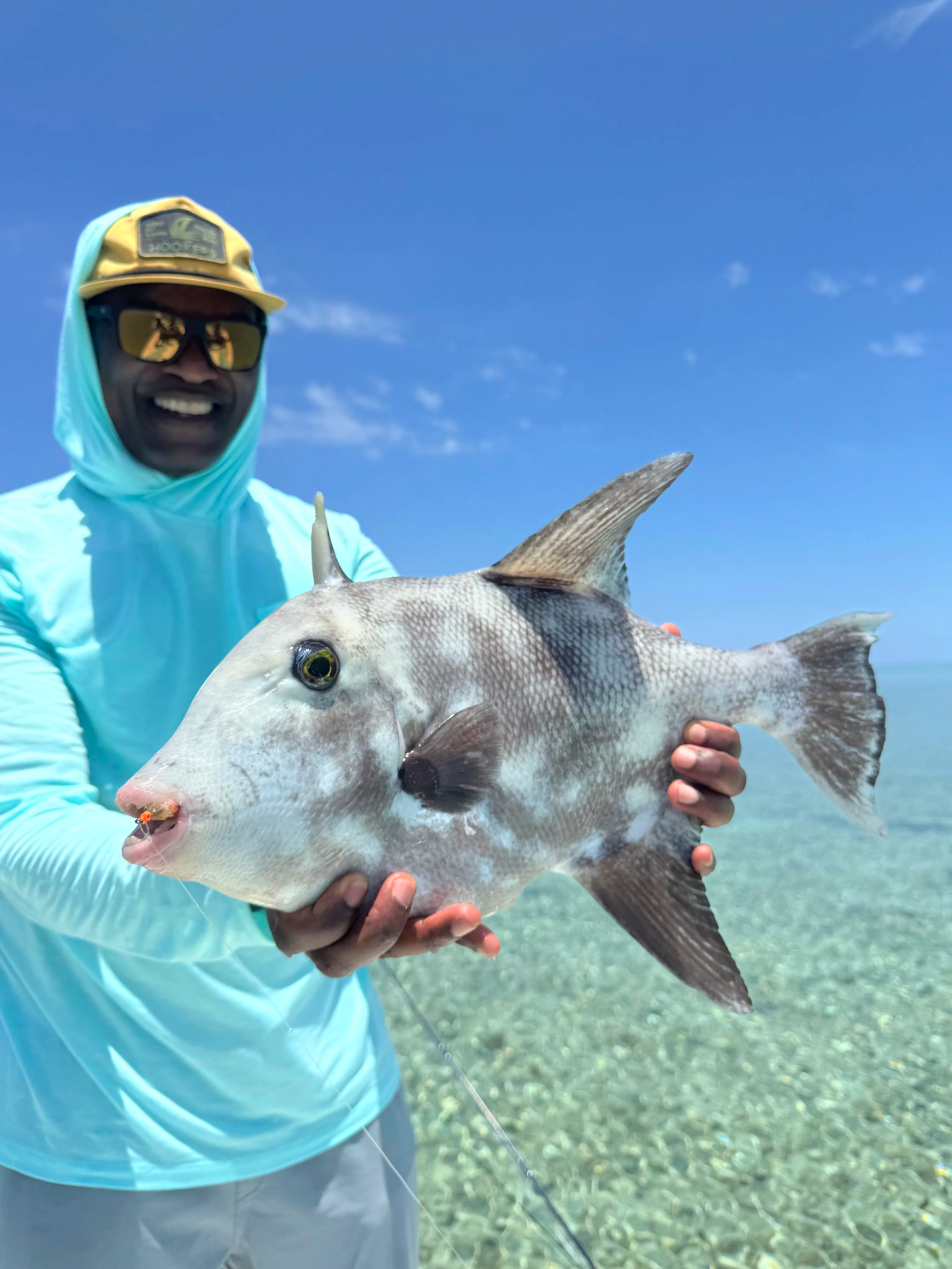 A man wearing a yellow cap, sunglasses, and a light blue shirt holding a large fish with a grey body and black eyes at the beach during daytime.