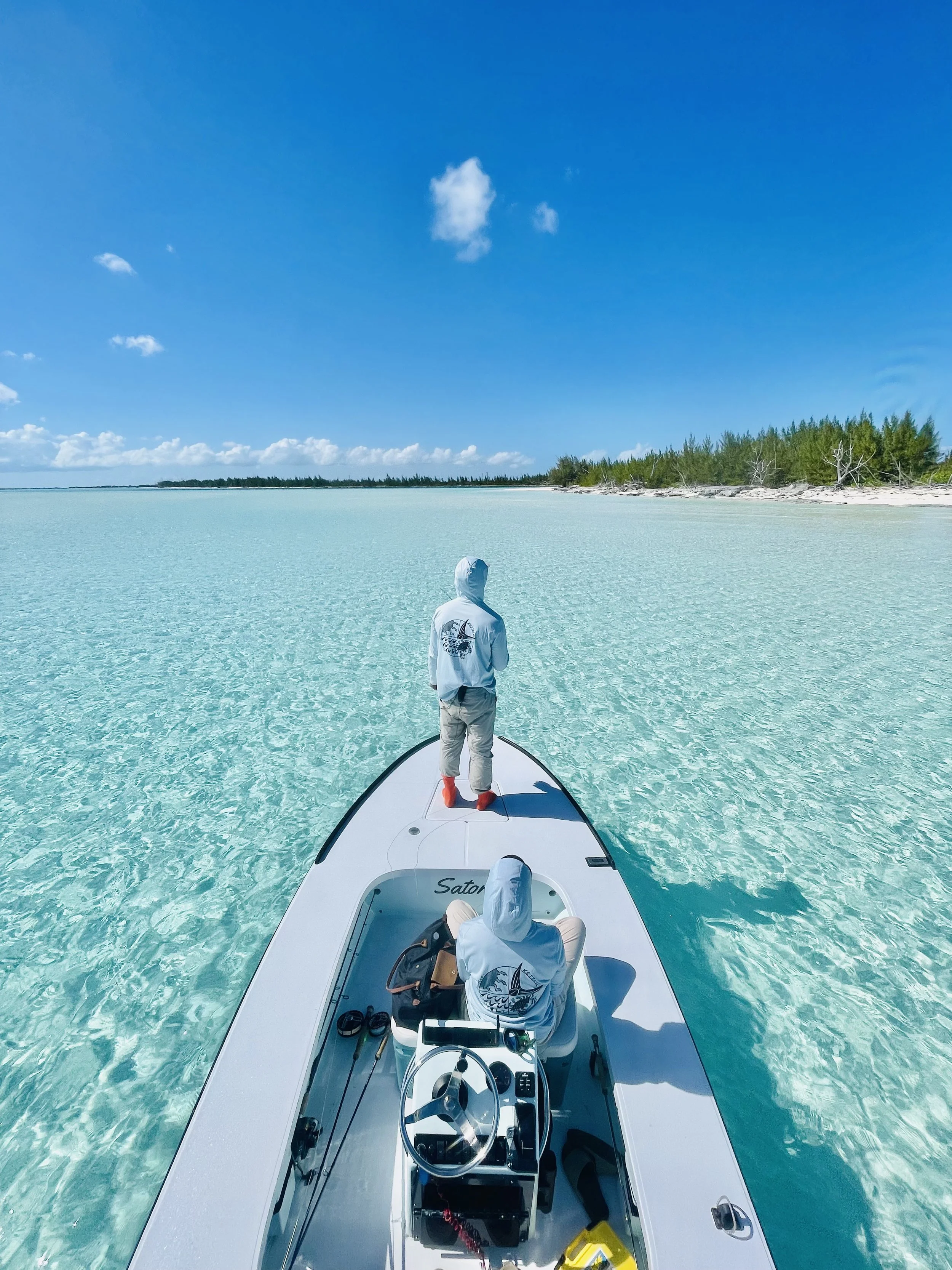 Person standing at the bow of a boat in clear turquoise water near a shoreline with green trees under a blue sky with a few clouds.