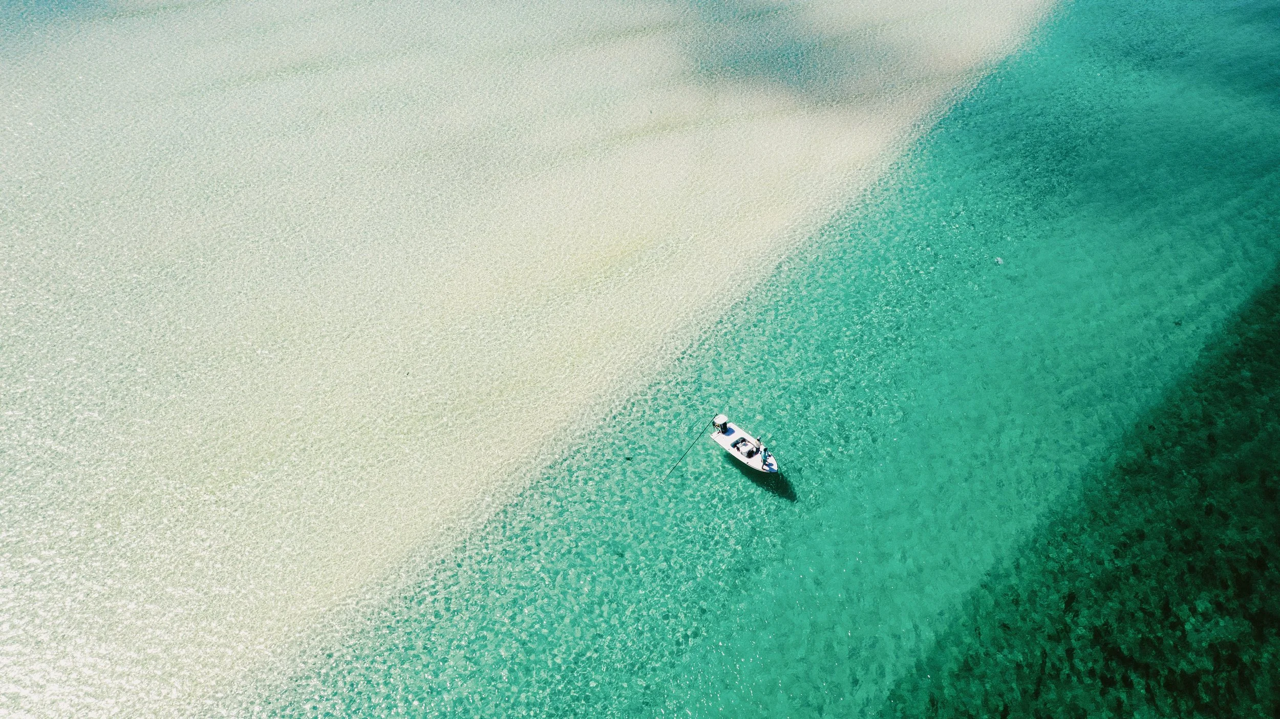 Aerial view of a flats skiff poling through clear turquoise water and a white-sand flat in Grand Bahama, Bahamas.