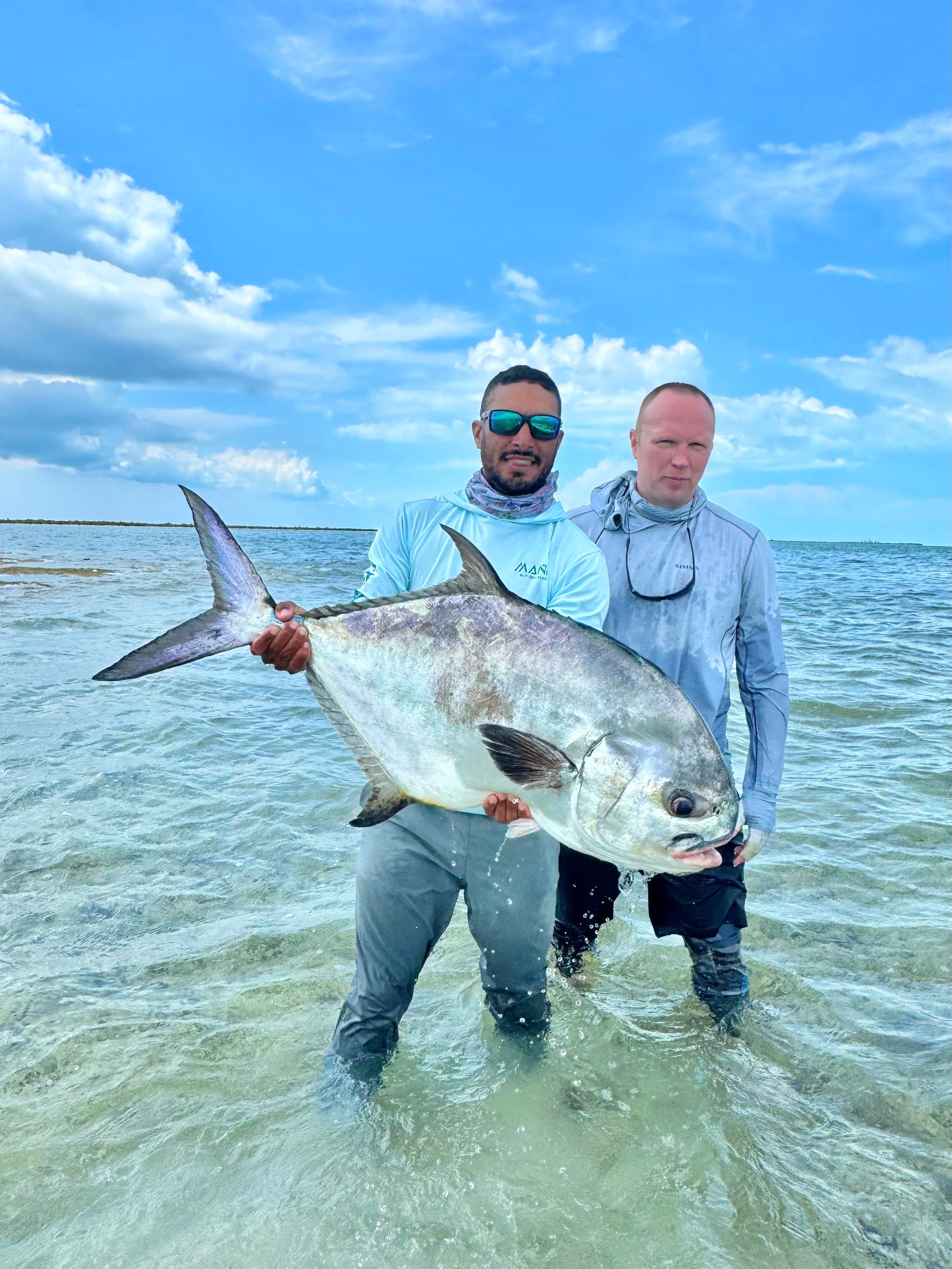 Two men standing in shallow water holding a large fish, with a blue sky and clouds overhead.