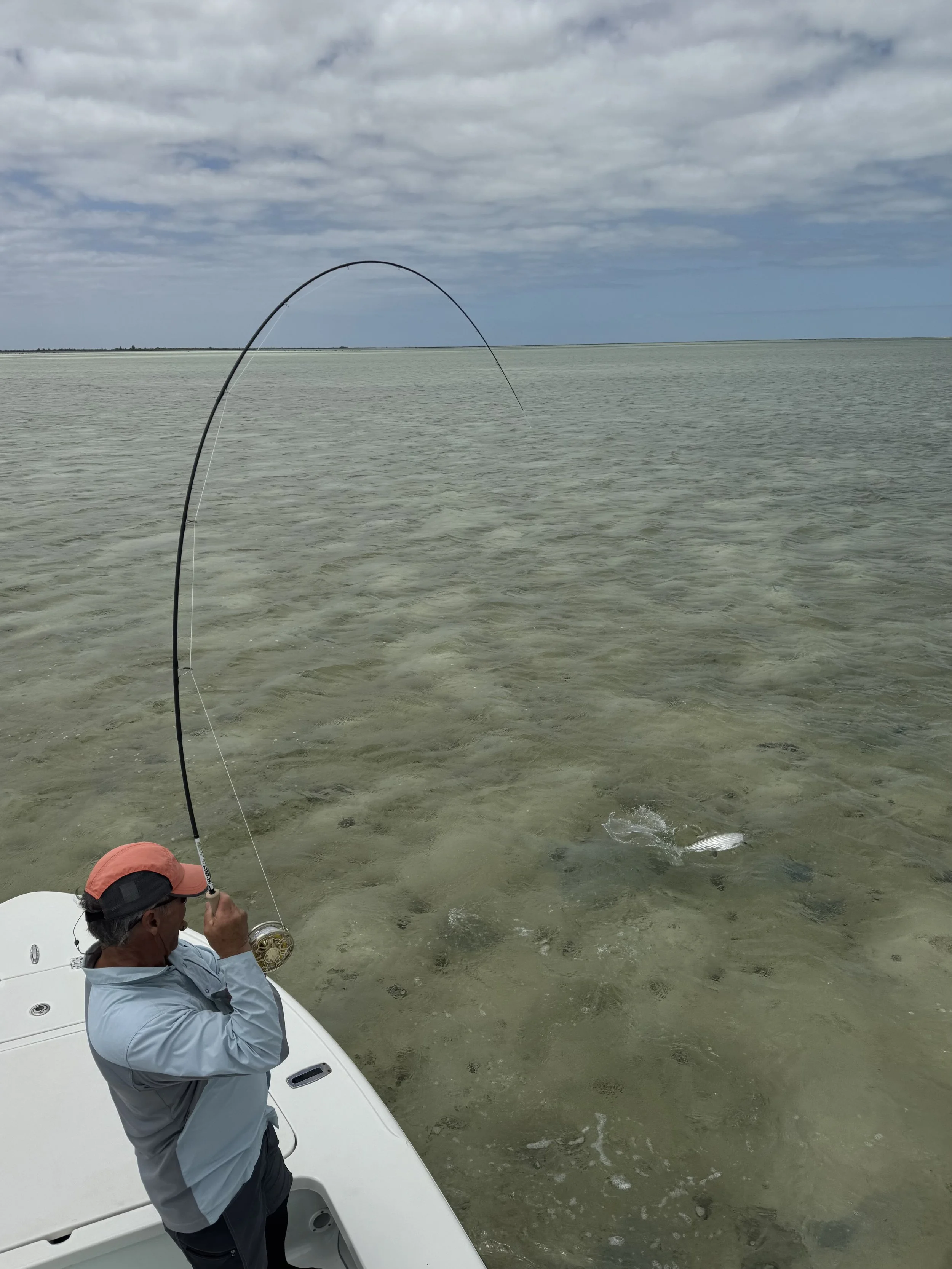 A person fishing on a boat in shallow water with a fishing rod, wearing a hat and a light blue jacket, under a cloudy sky.