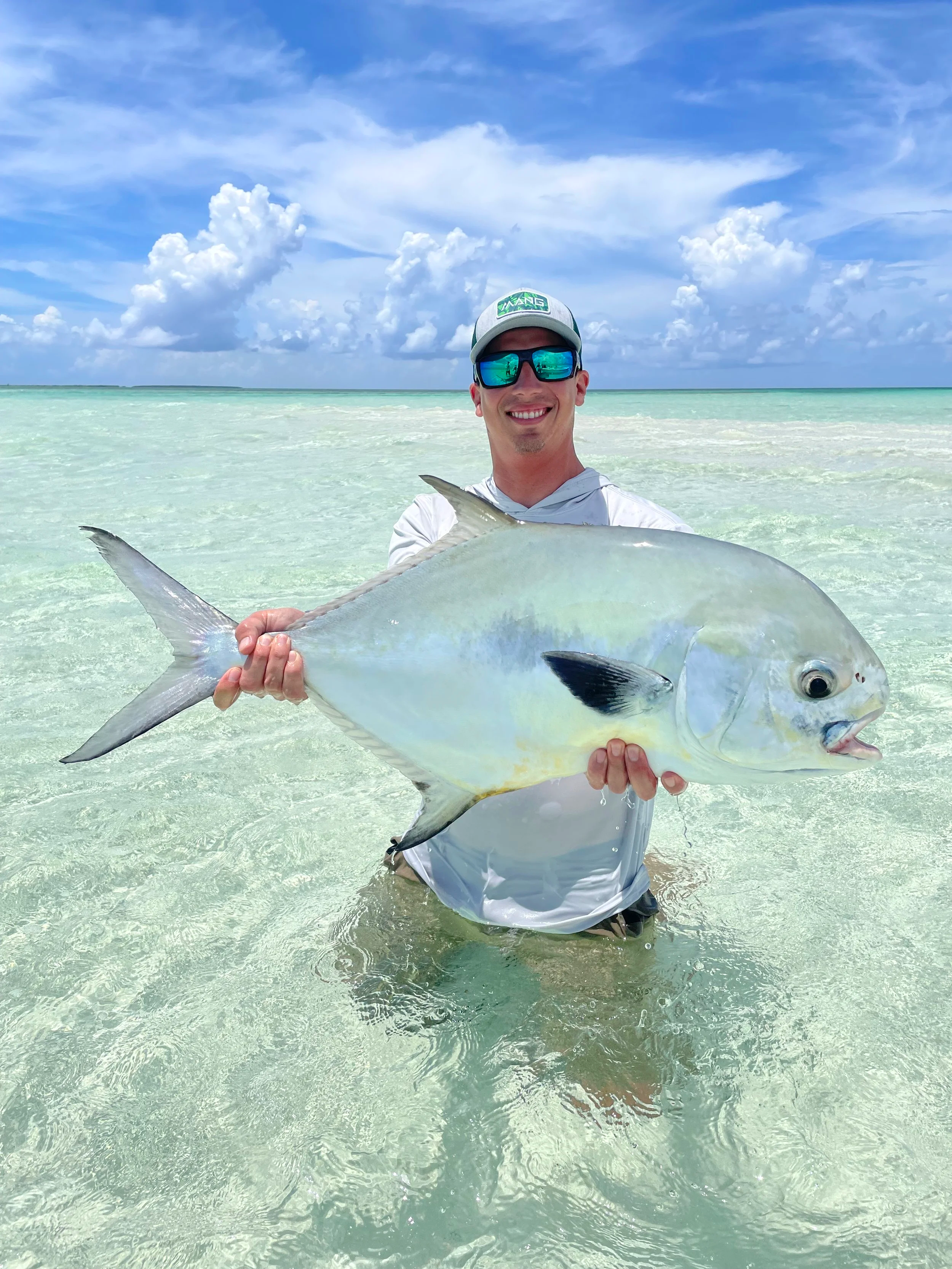 A man wearing sunglasses and a cap is standing in shallow, clear water on a beach, holding a large fish.