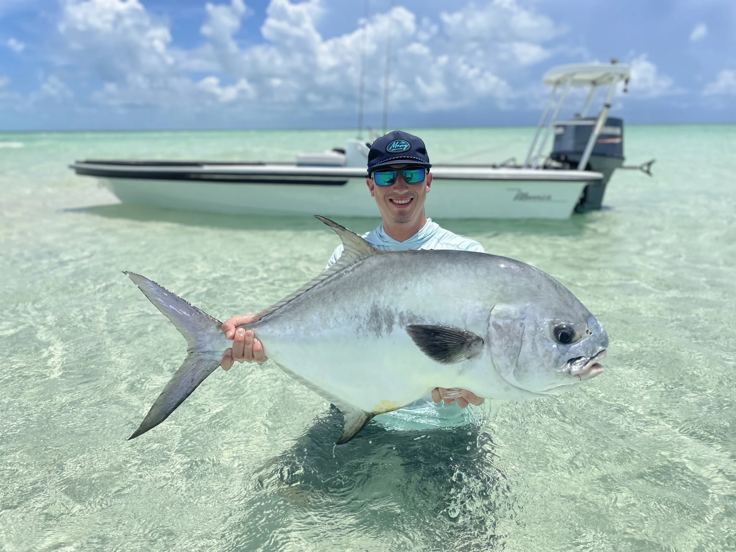 A man wearing sunglasses and a cap is smiling and holding a large fish in shallow, clear water with a boat in the background under partly cloudy skies.