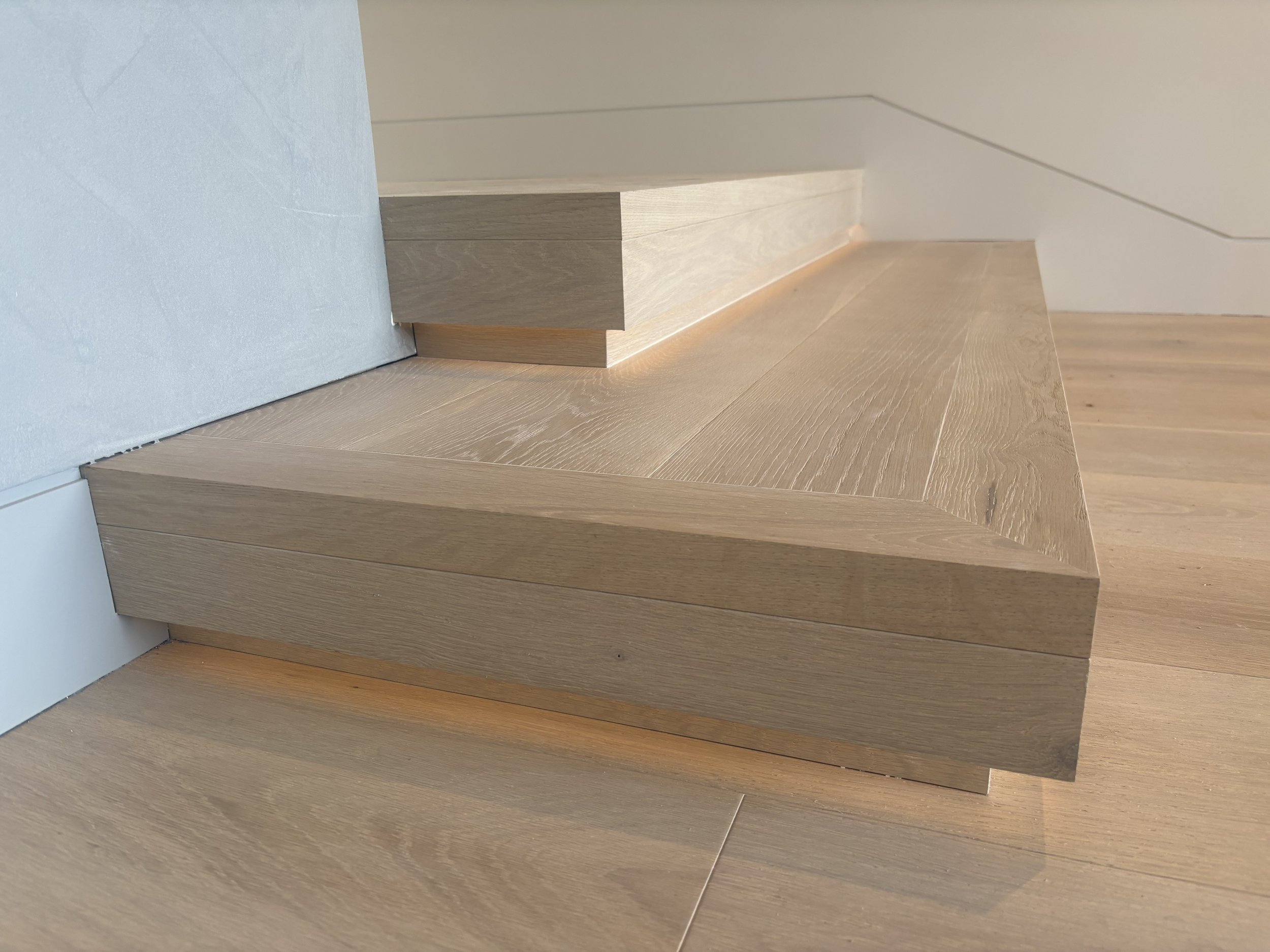Close-up of wooden stairs and flooring, with a light-colored wall and shadowed area.