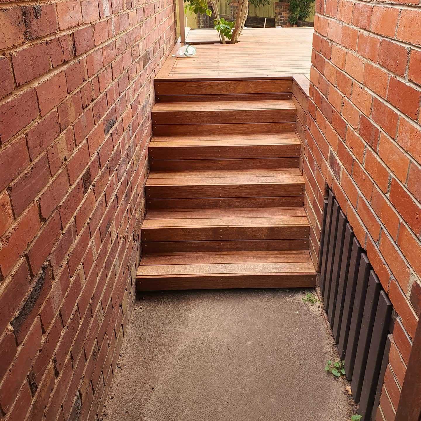 A small outdoor area with wooden stairs connecting a lower concrete patio to an upper wooden deck. The area is bordered by red brick walls, with some green plants visible in the background.