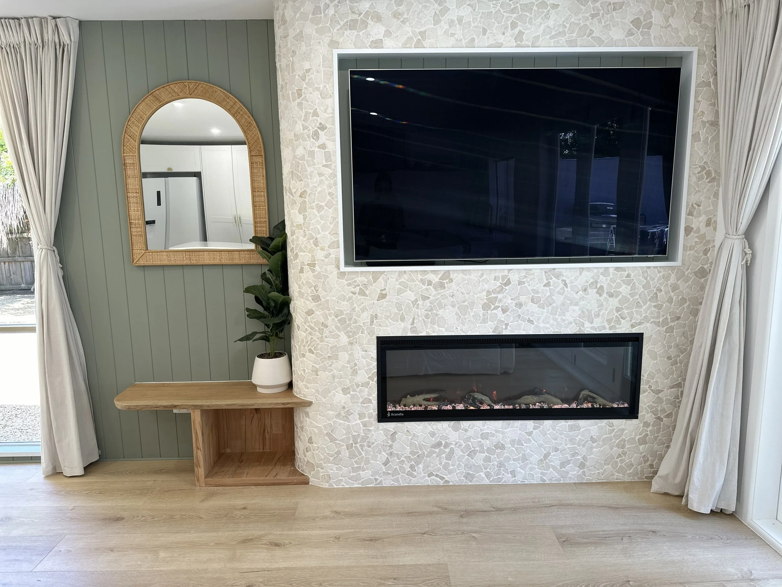 Living room corner with a flat-screen TV mounted on a stone accent wall, a small wooden table, a potted plant, a mirror, and white curtains.