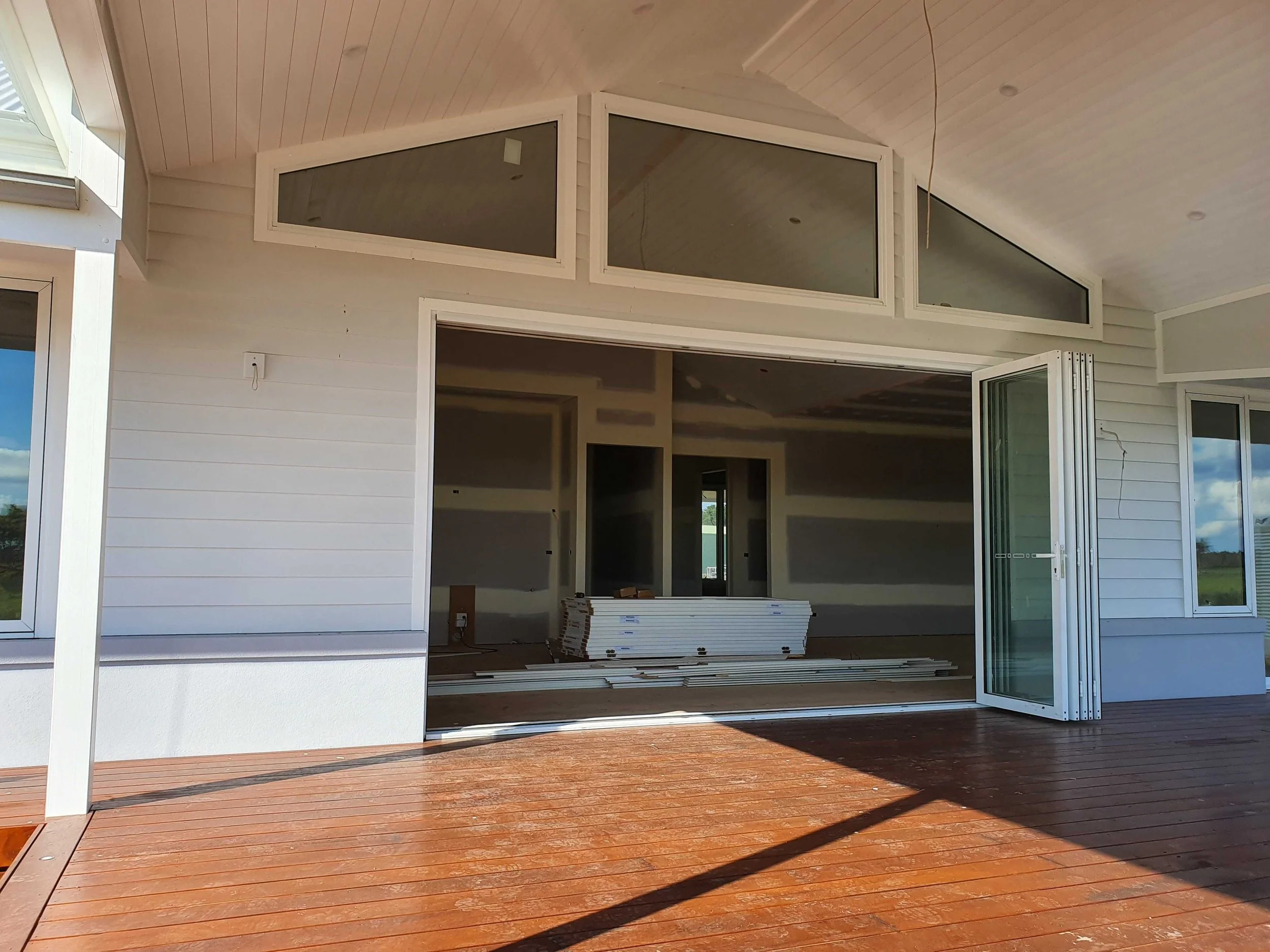 The image shows the exterior of a house under construction with an open sliding glass door leading to a wooden deck. The interior of the house is visible, with construction materials and an open wall.