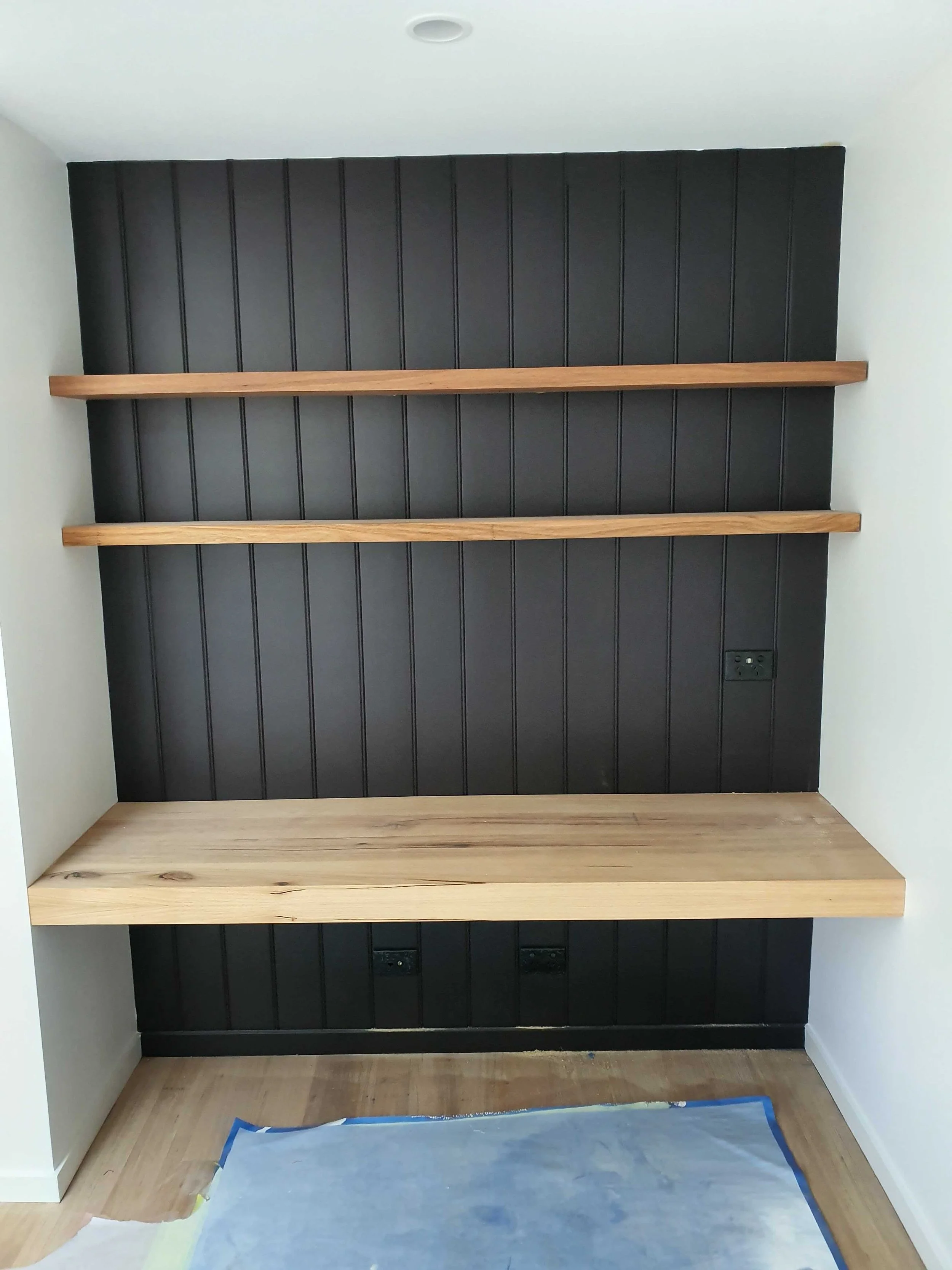 A partially completed built-in wall unit with black vertical paneling, three wooden shelves, and a light wood base, against a white wall with a ceiling recess, and an electrical outlet near the bottom right.