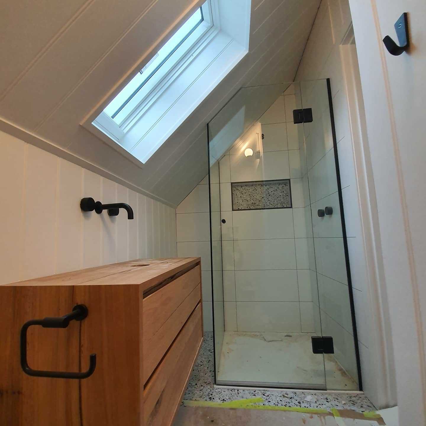 Interior of a bathroom under construction with a glass shower enclosure, white tiled walls, a wooden vanity, and a window with a skylight.