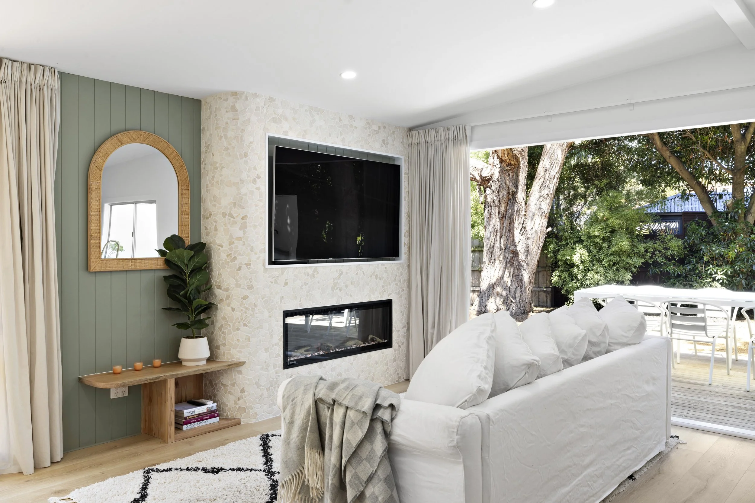 Living room with a large white sofa, a wall-mounted flat-screen TV, a fireplace, a mirror with a wooden frame, and large windows showing outdoor trees and patio furniture.