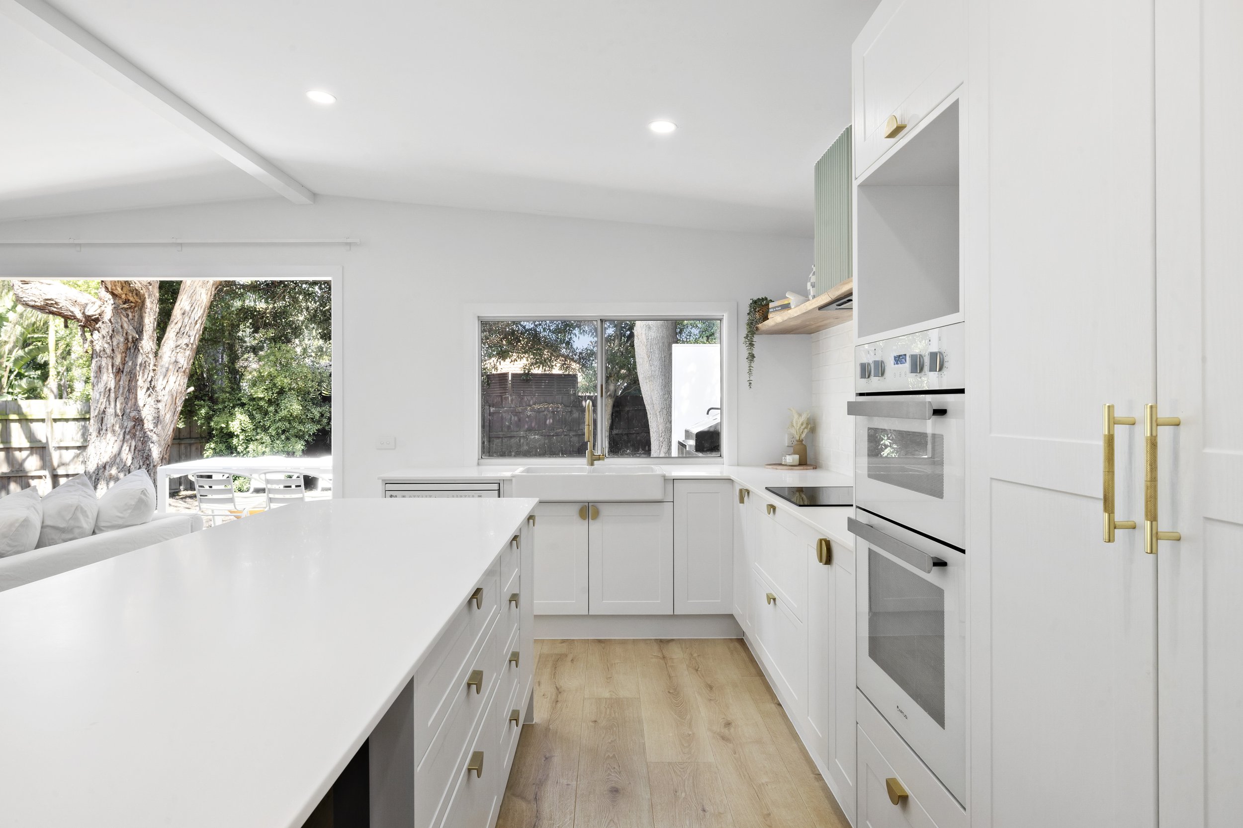 Modern white kitchen with gold hardware, wooden floor, and large windows overlooking a backyard with trees.