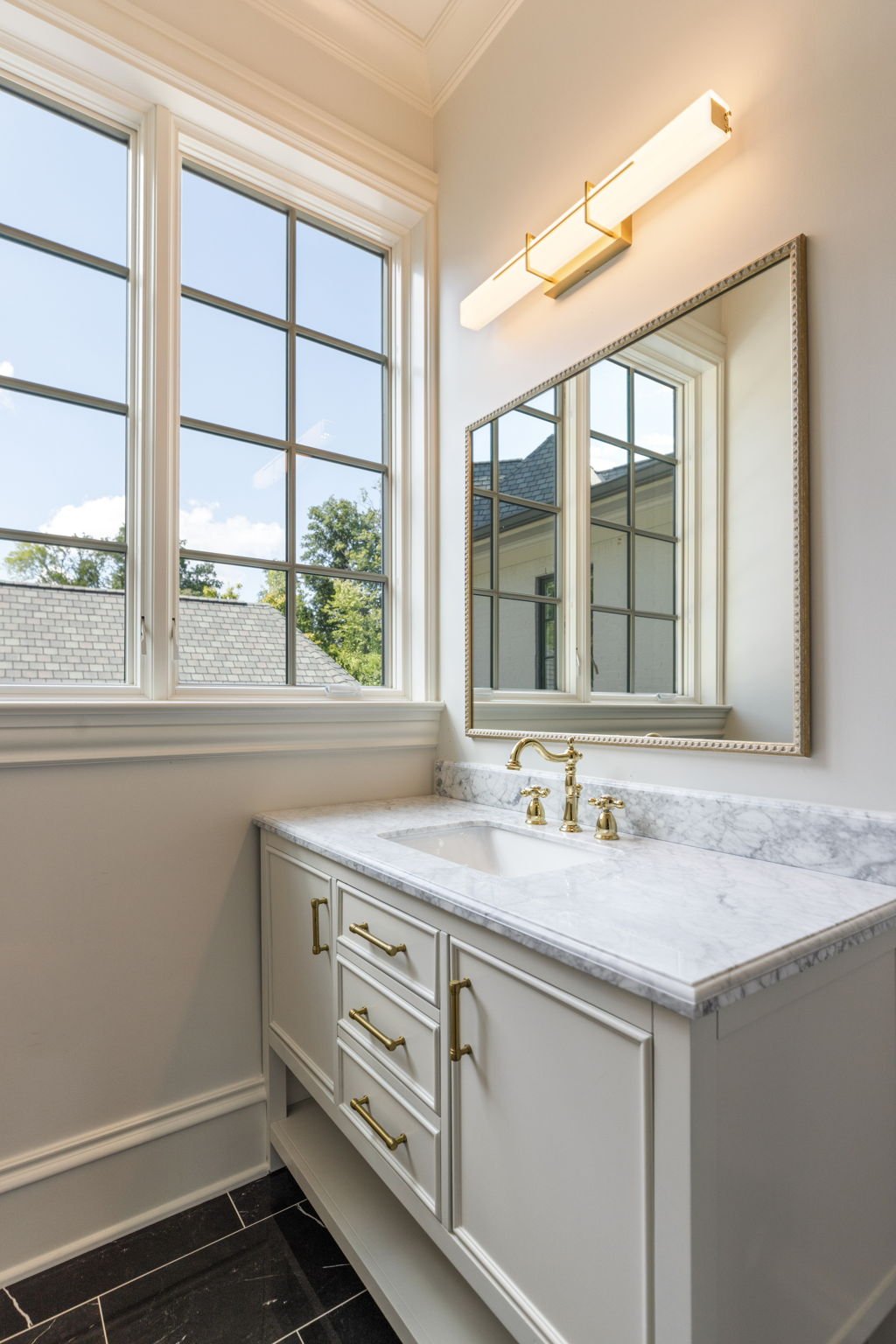 Bathroom vanity with white cabinetry, marble countertop, and gold fixtures. Large window with grid panes reflecting in a mirror above the sink, and modern wall-mounted light fixture.
