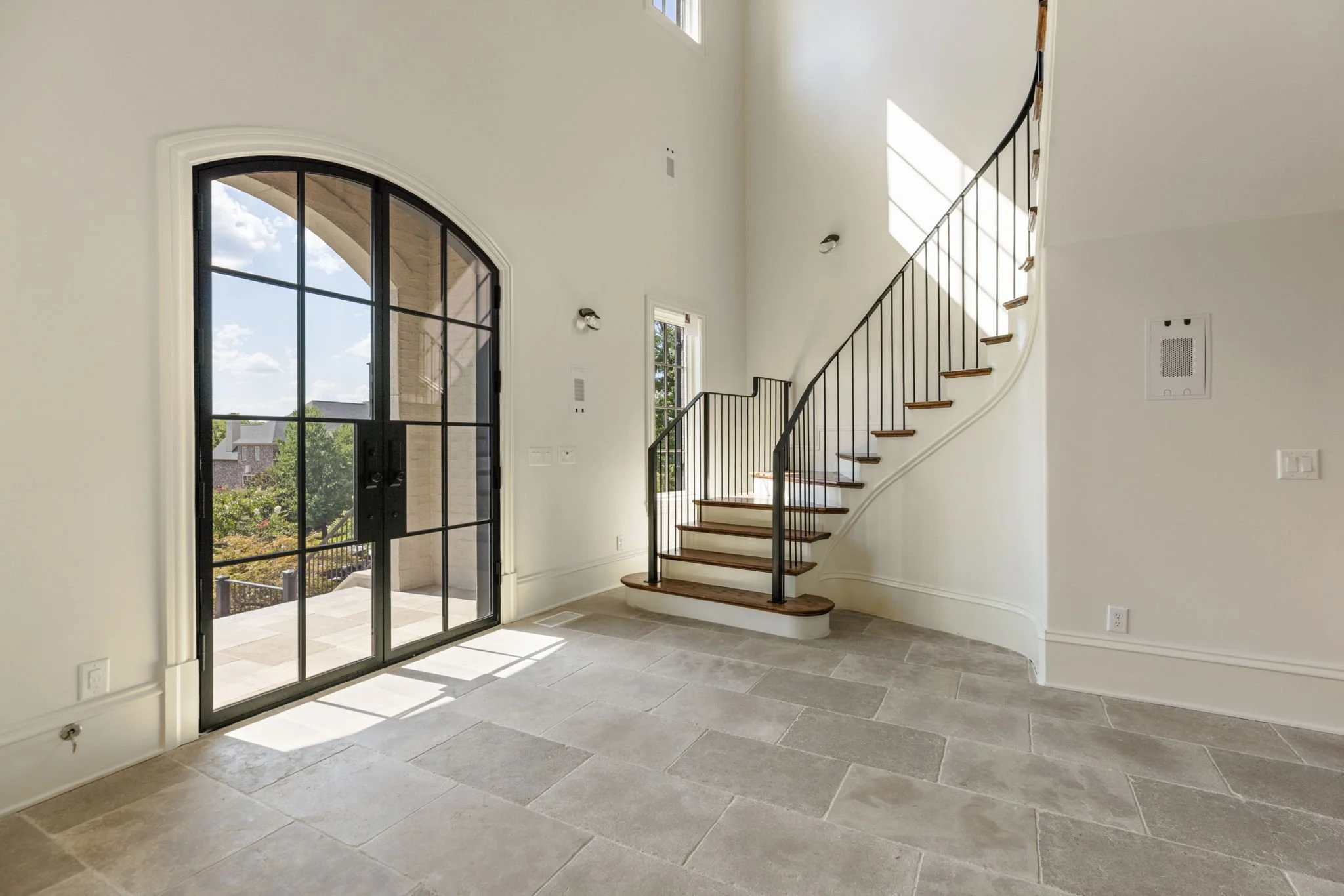 Interior view of a modern house entryway with a staircase, large black-framed arched window, and natural sunlight.