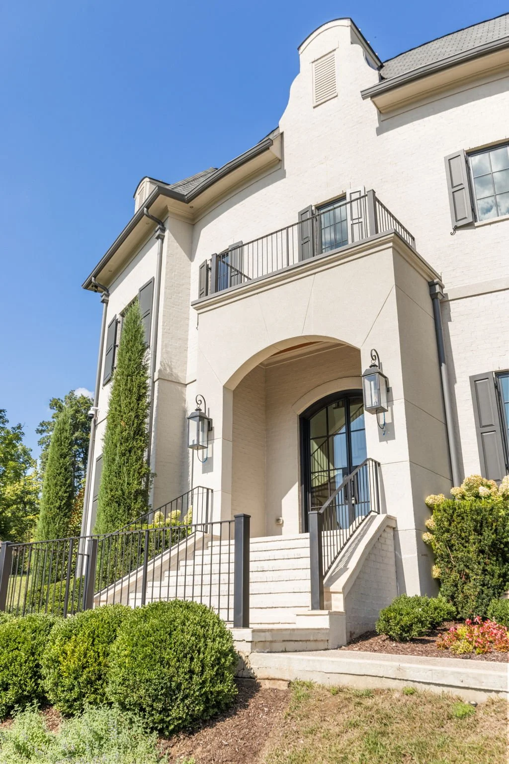 Front exterior of a white multi-story house with black shutters, a staircase with black railing, and a small balcony with a black railing under a clear blue sky.