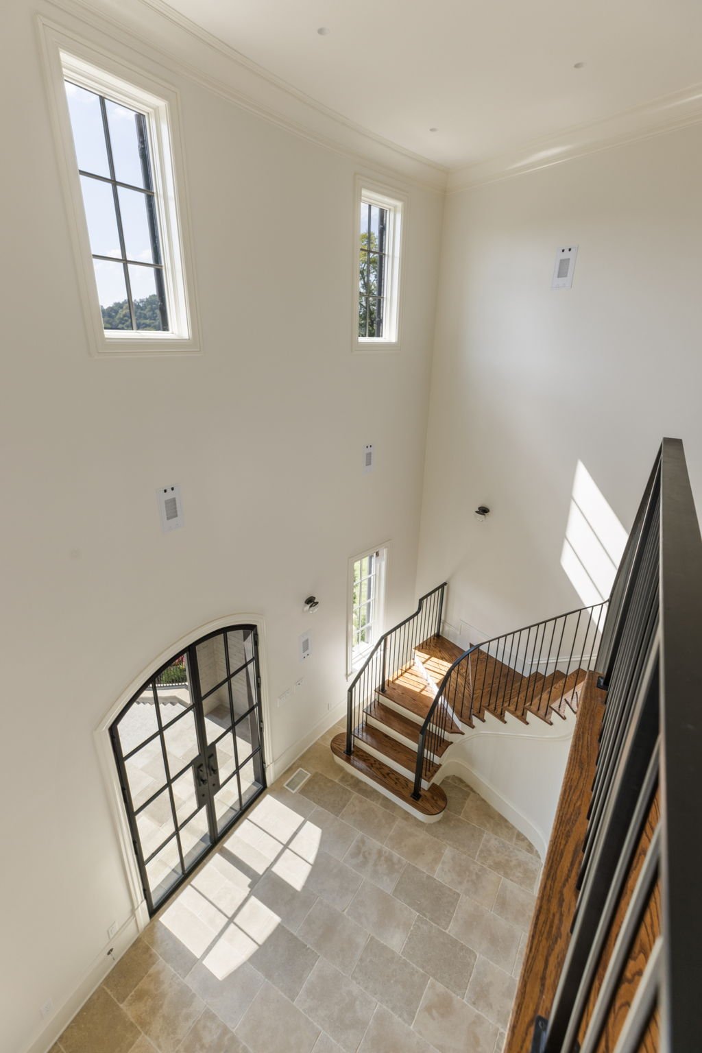 A bright, spacious foyer with high ceiling, large windows, a wooden staircase with black metal railing, limestone floor, and a glass double door leading outside.