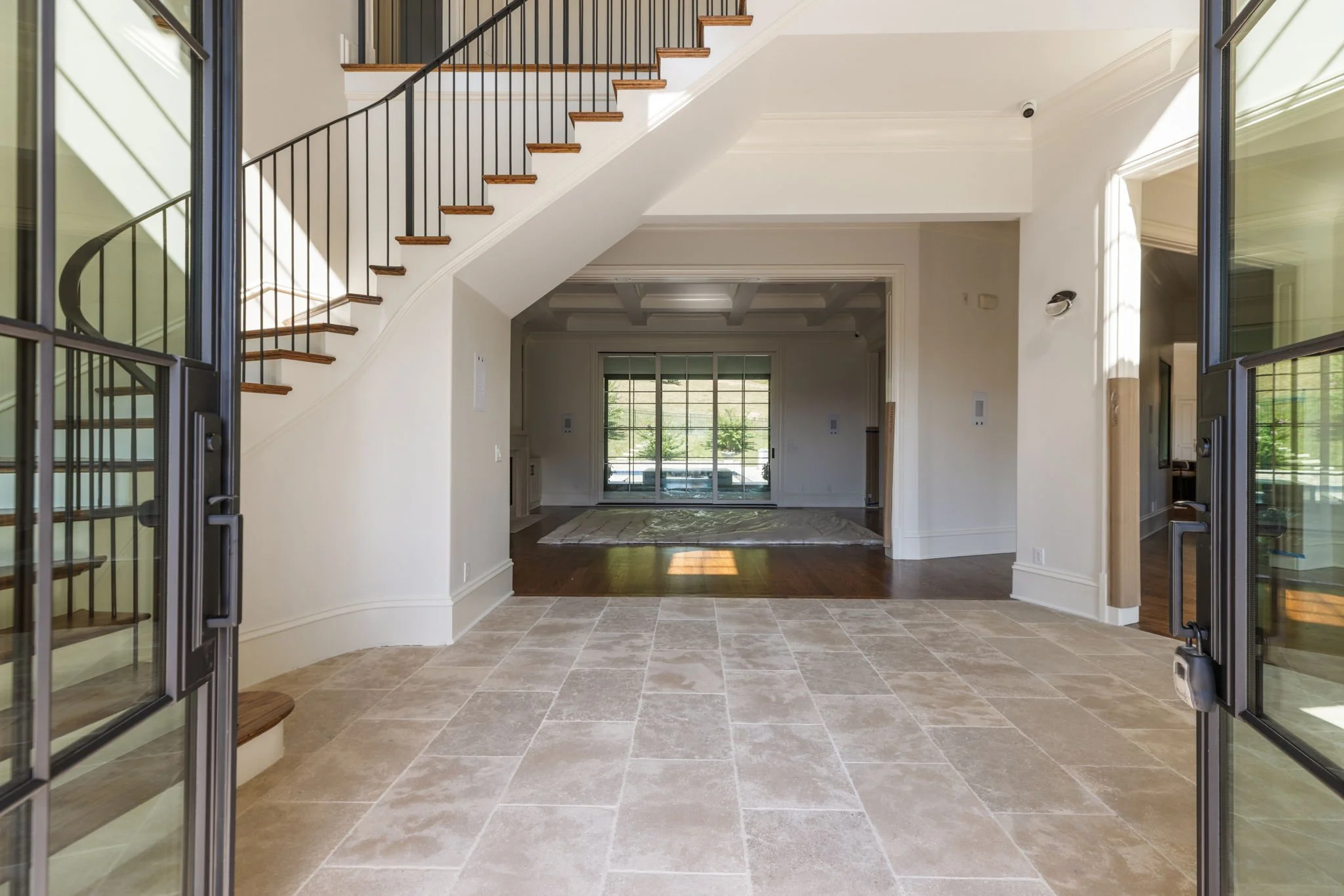 View of a spacious, modern home interior with a staircase having wooden steps and black metal railing, leading to an upper floor. The entry area features tile flooring and opens into a living space with a large window and sliding glass door showing o
