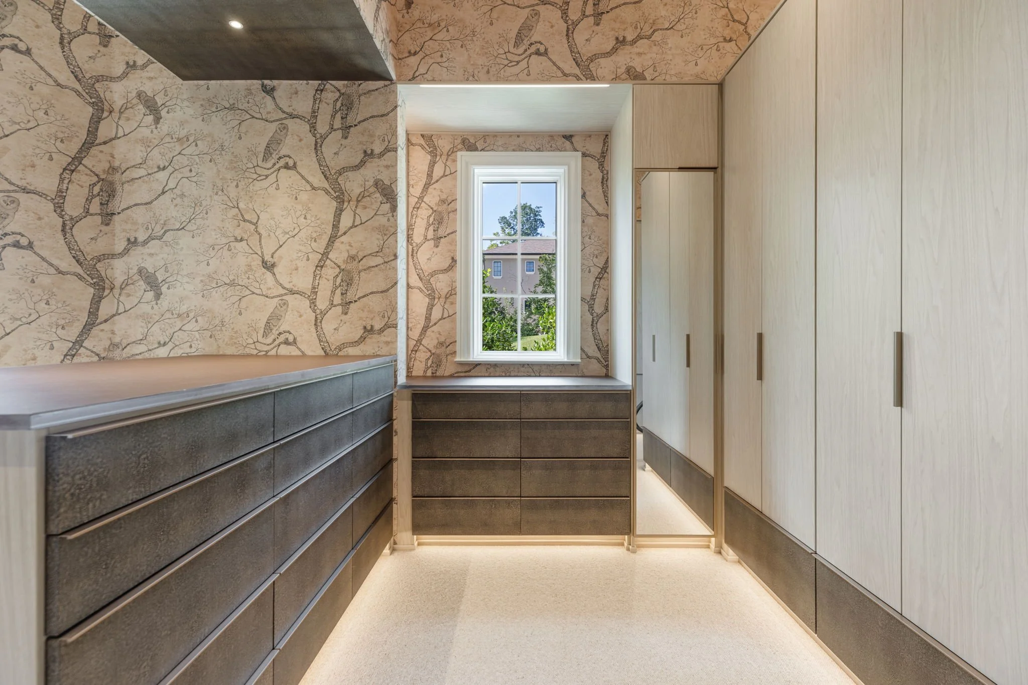 A walk-in closet with light wood cabinets on the right and dark gray drawers on the left, a single window allowing natural light, and wallpaper with a tree and owl pattern on the walls and ceiling.