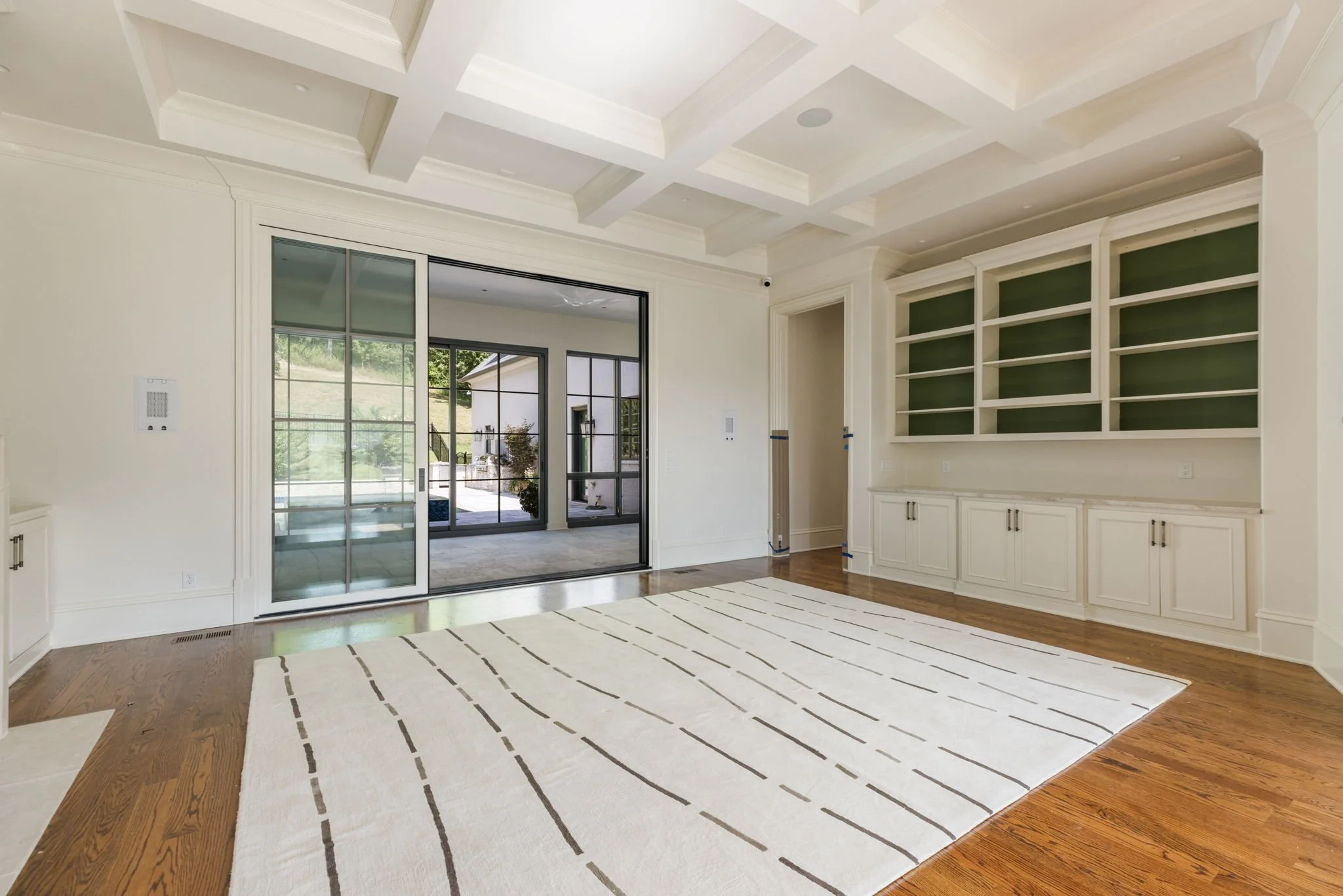 Bright living room with wood floor, white walls, built-in bookshelves with green backing, large area rug, and sliding glass doors leading to the sun room.